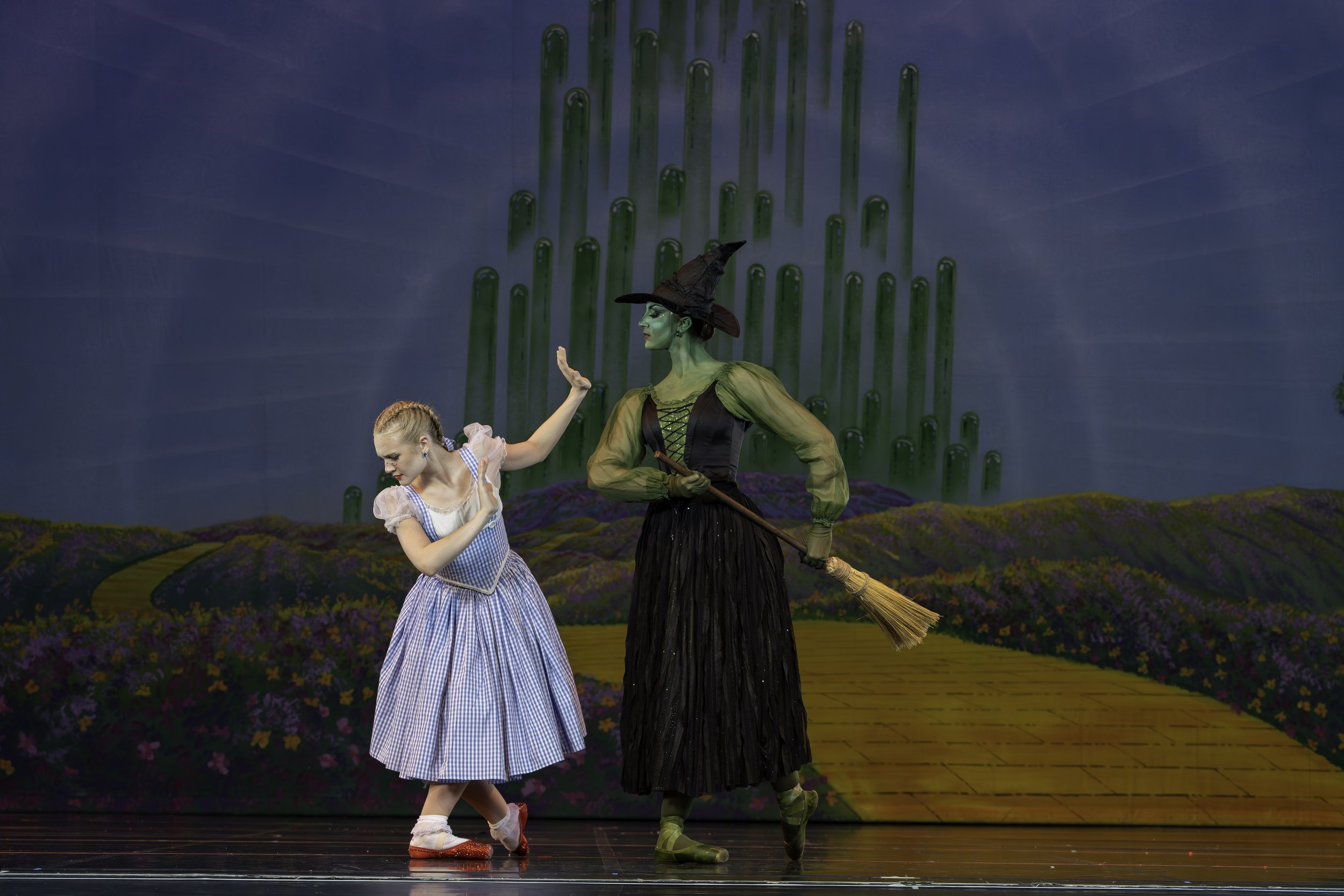 A girl dressed as Dorothy from The Wizard of Oz dancing with the Wicked Witch of the West, holding a broomstick, on a stage with a colorful painted backdrop of a yellow brick road and green tornado.