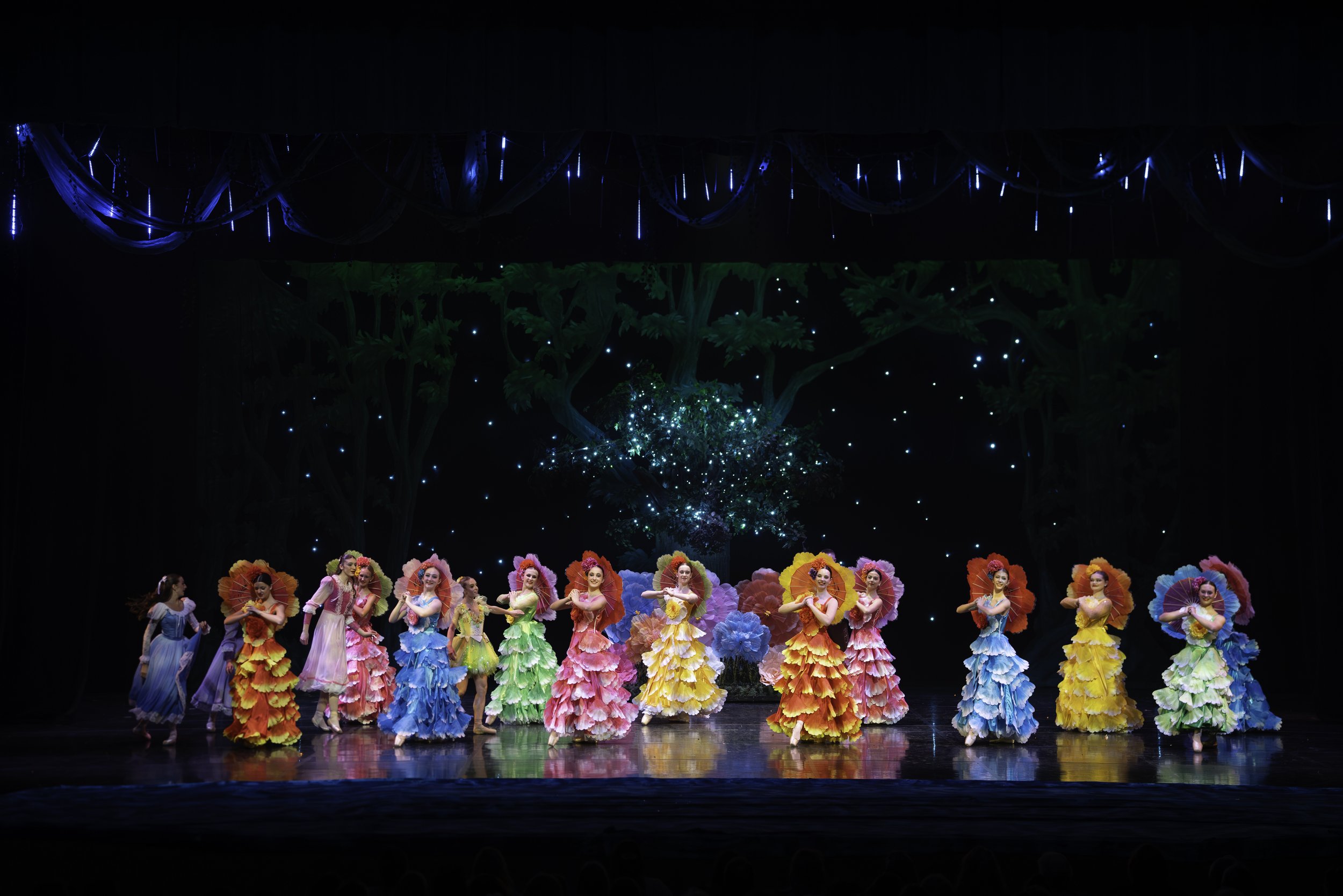 Colorful group of dancers wearing vibrant, floral-themed costumes performing on a stage with a backdrop of trees and a starry sky.