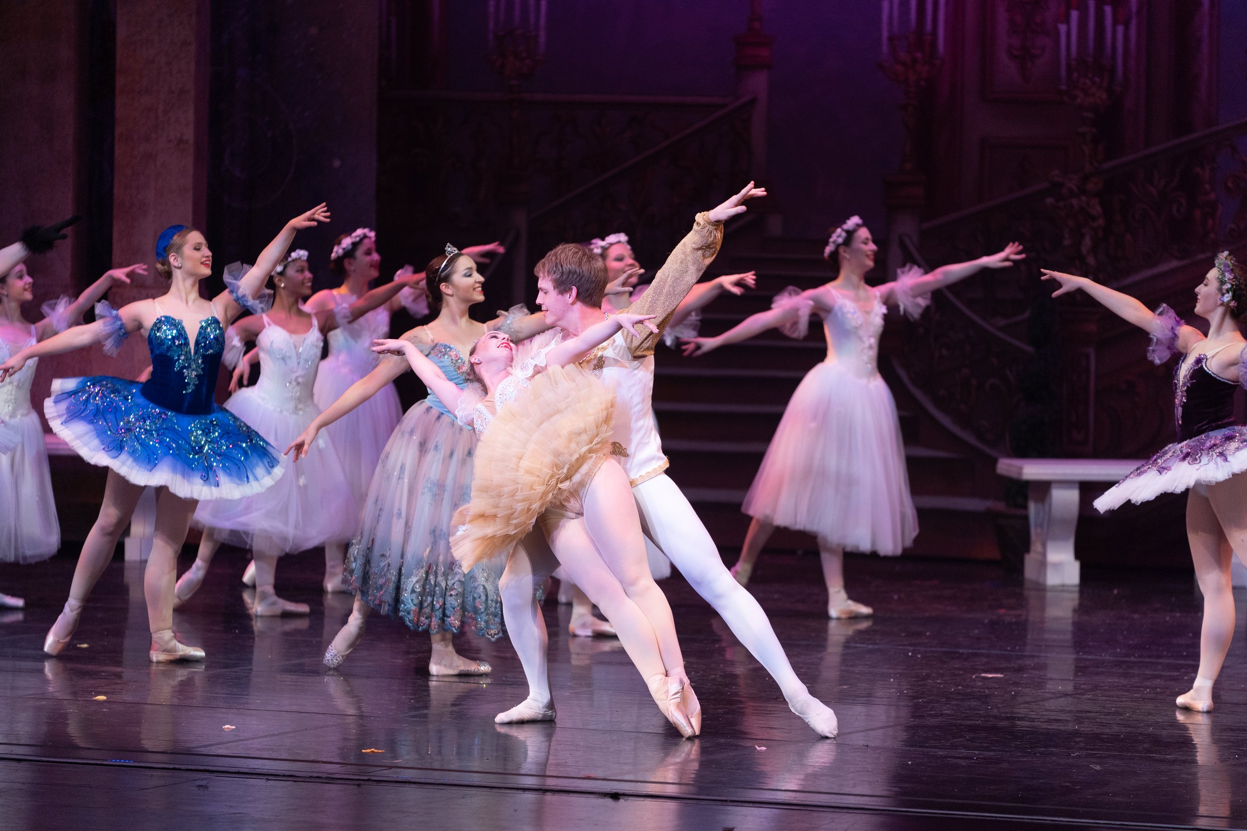 Ballet dancers performing on stage, with a male dancer holding a female dancer in a pose. Other dancers in tutus and ballet costumes participate in the performance.
