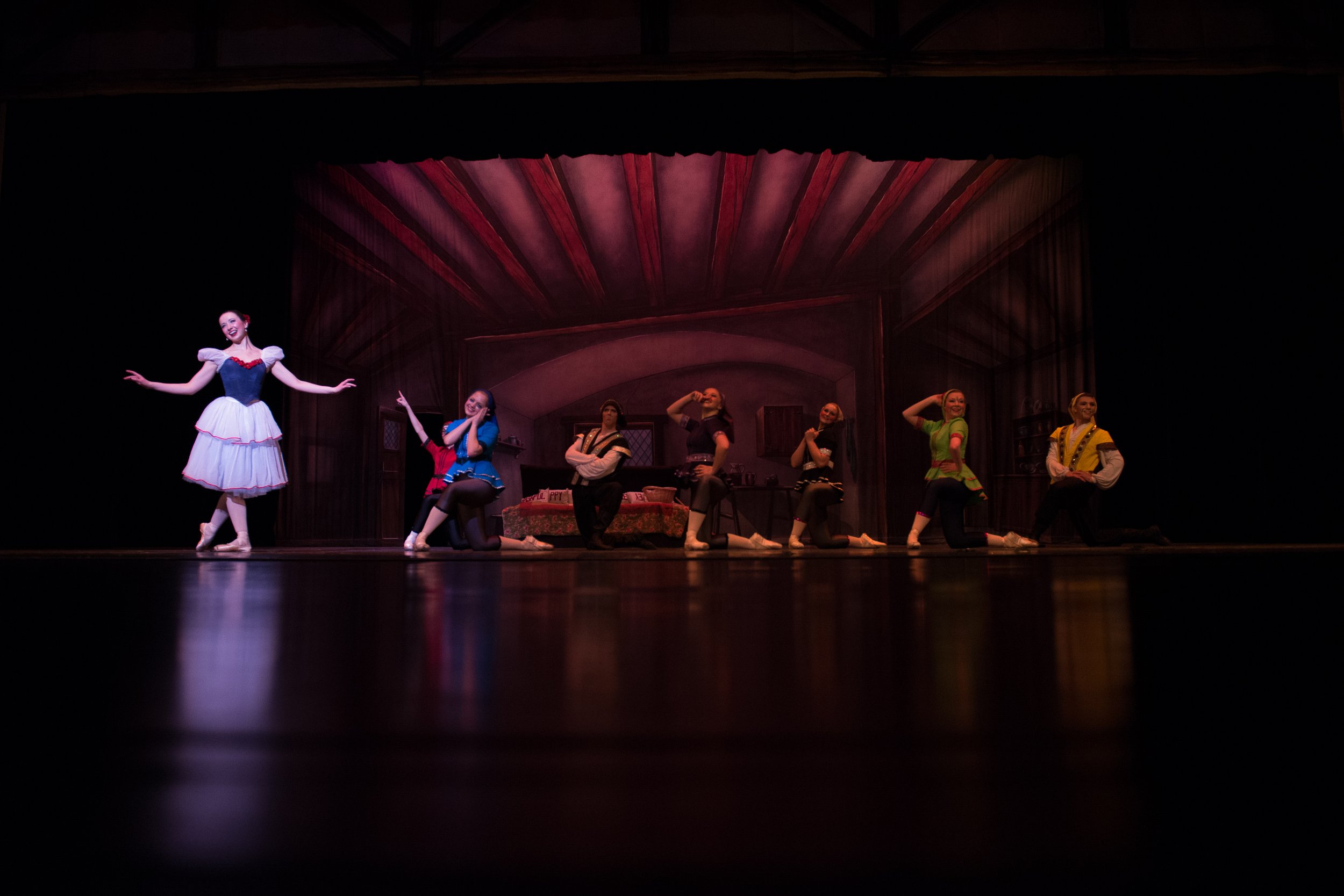 Theatrical stage scene with eight performers in colorful costumes, some kneeling and others standing, set against a detailed painted backdrop of a rustic interior. One performer stands apart, smiling with arms outstretched.