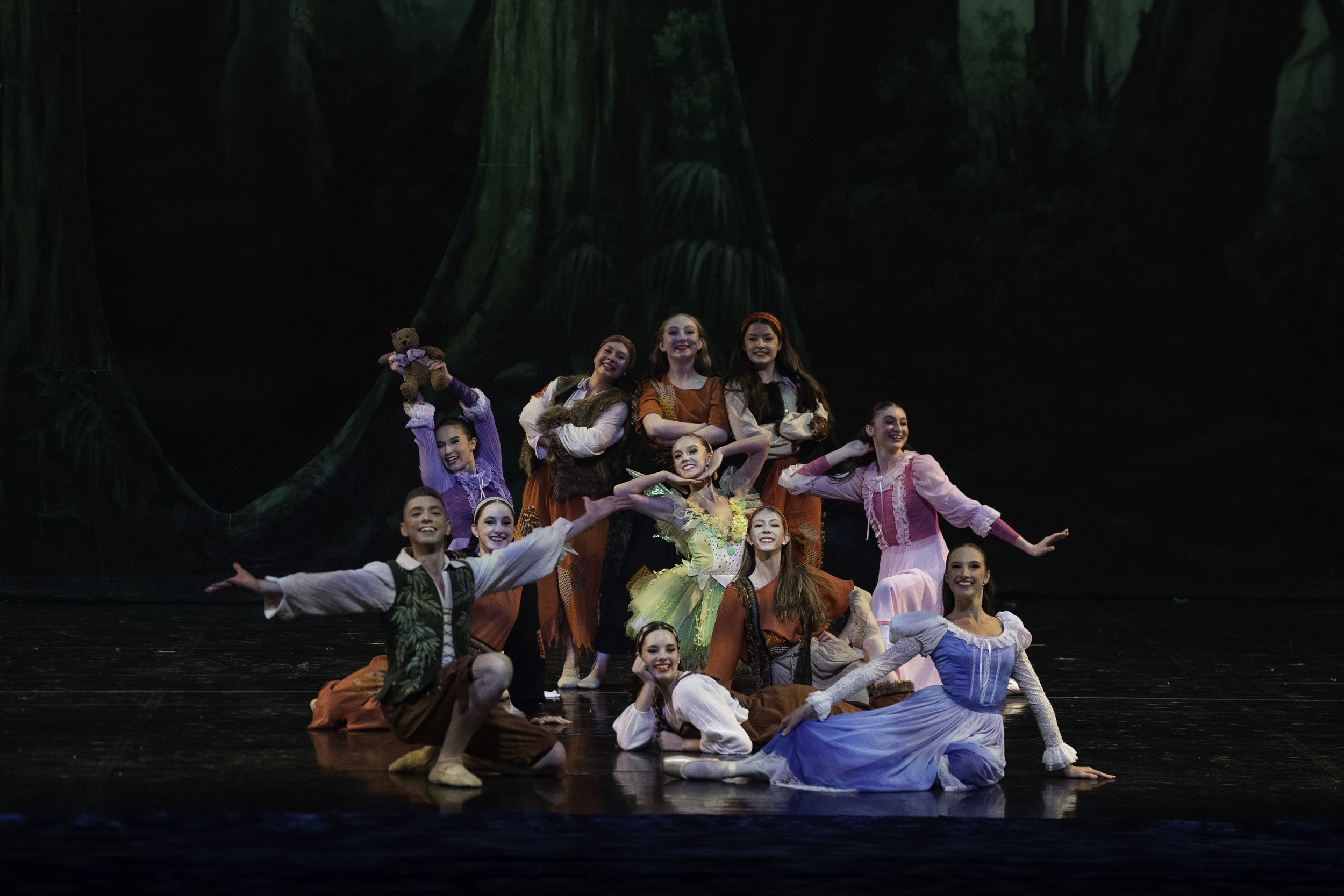 A group of young performers in colorful costumes on stage, posing for a cast photo during a dance or theater production, with a dark forest backdrop.