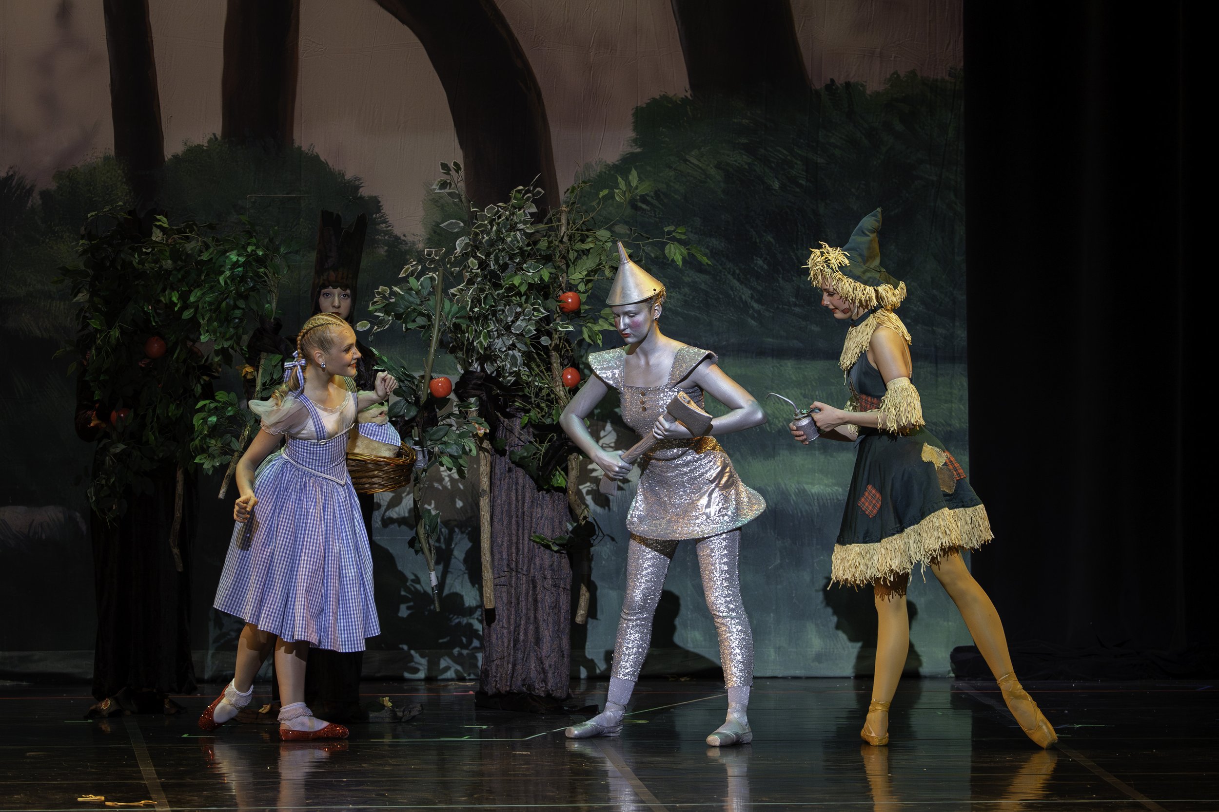 Children performing a theatrical play with characters dressed as characters from The Wizard of Oz, including Dorothy, the Tin Man, and the Scarecrow, on stage with a painted forest background.
