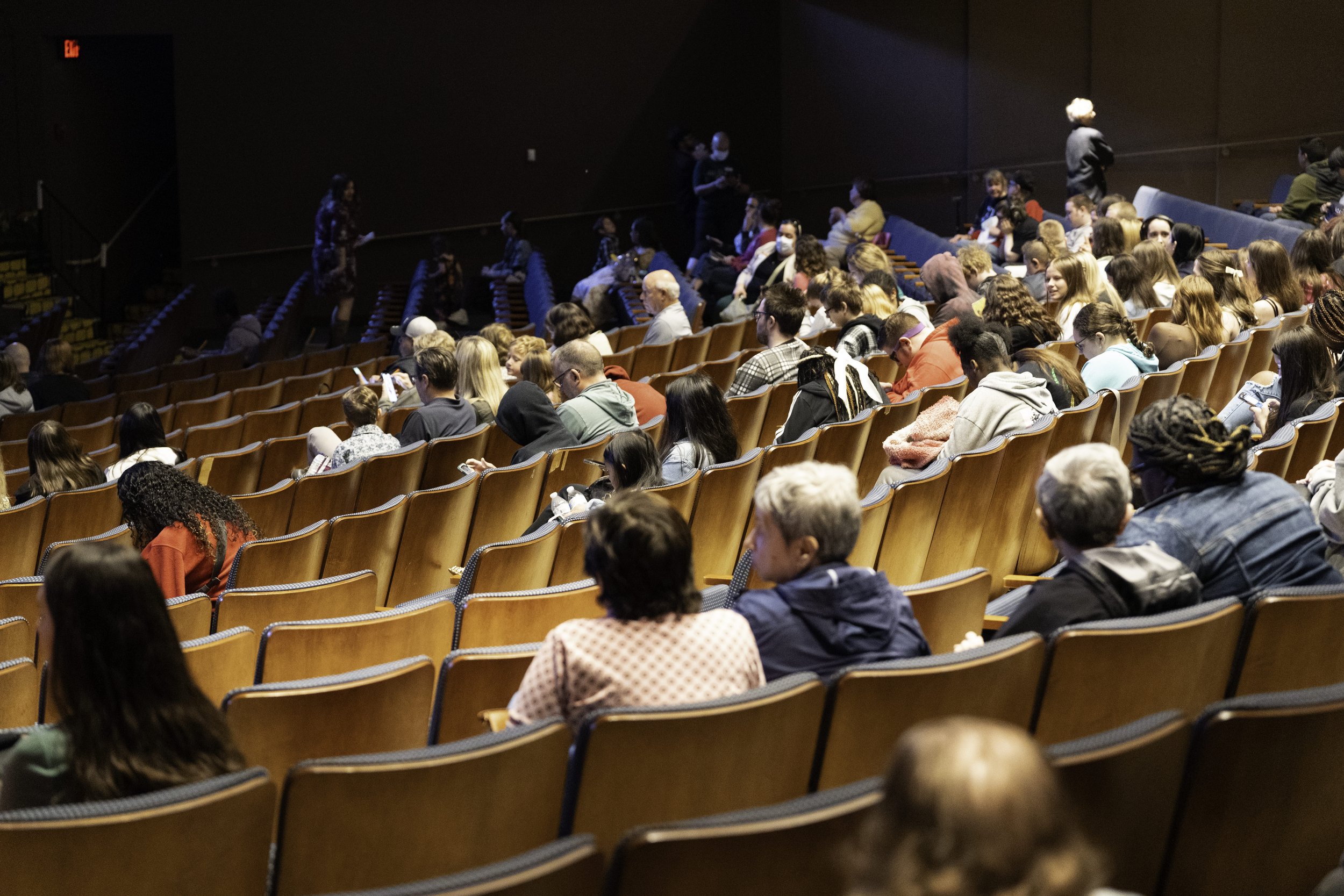 People seated in an auditorium, some using phones, and others talking or waiting for an event to start.