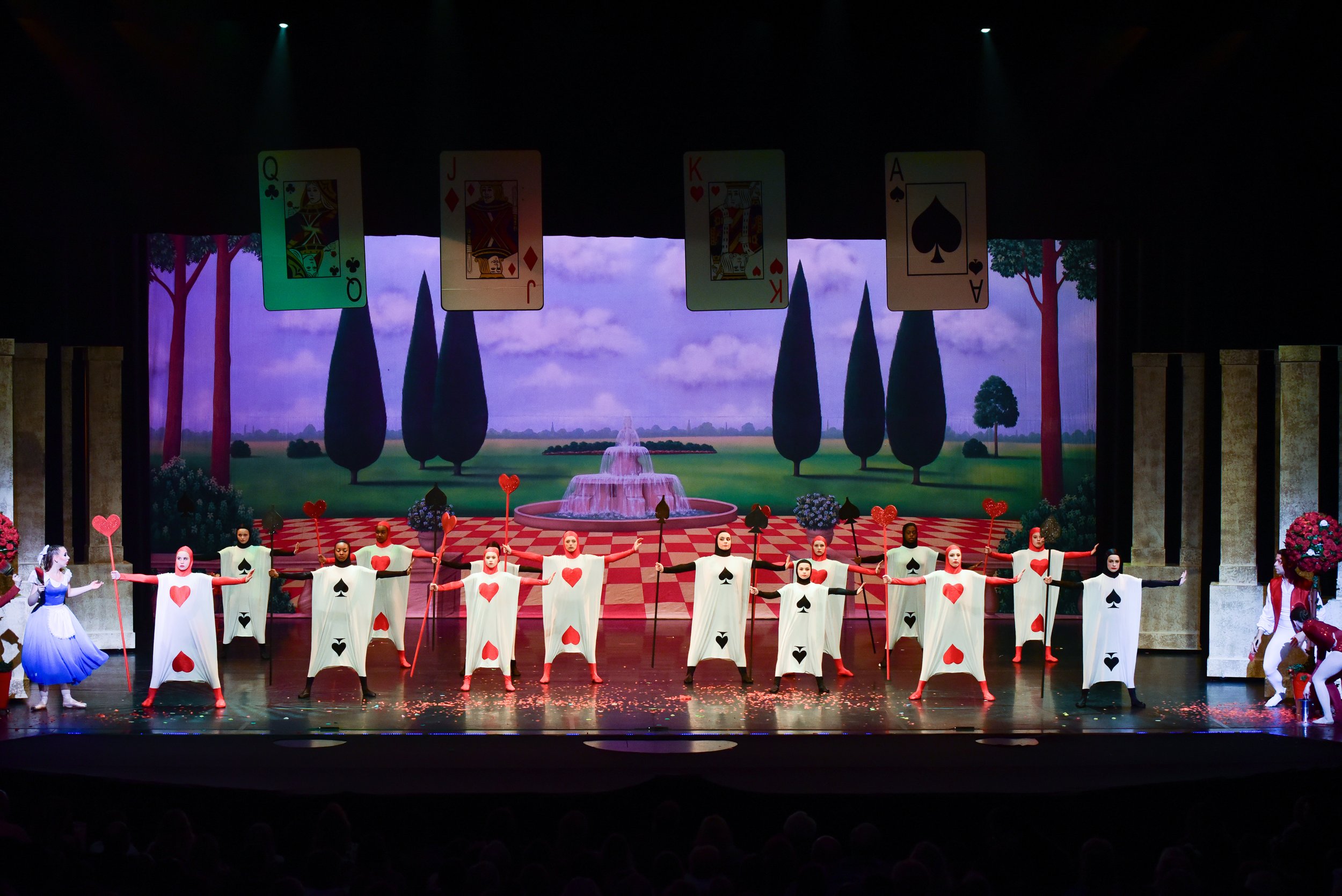 Theatrical stage performance of 'Alice in Wonderland' with dancers dressed as playing cards, set against a colorful backdrop featuring trees, a fountain, and playing cards overhead.