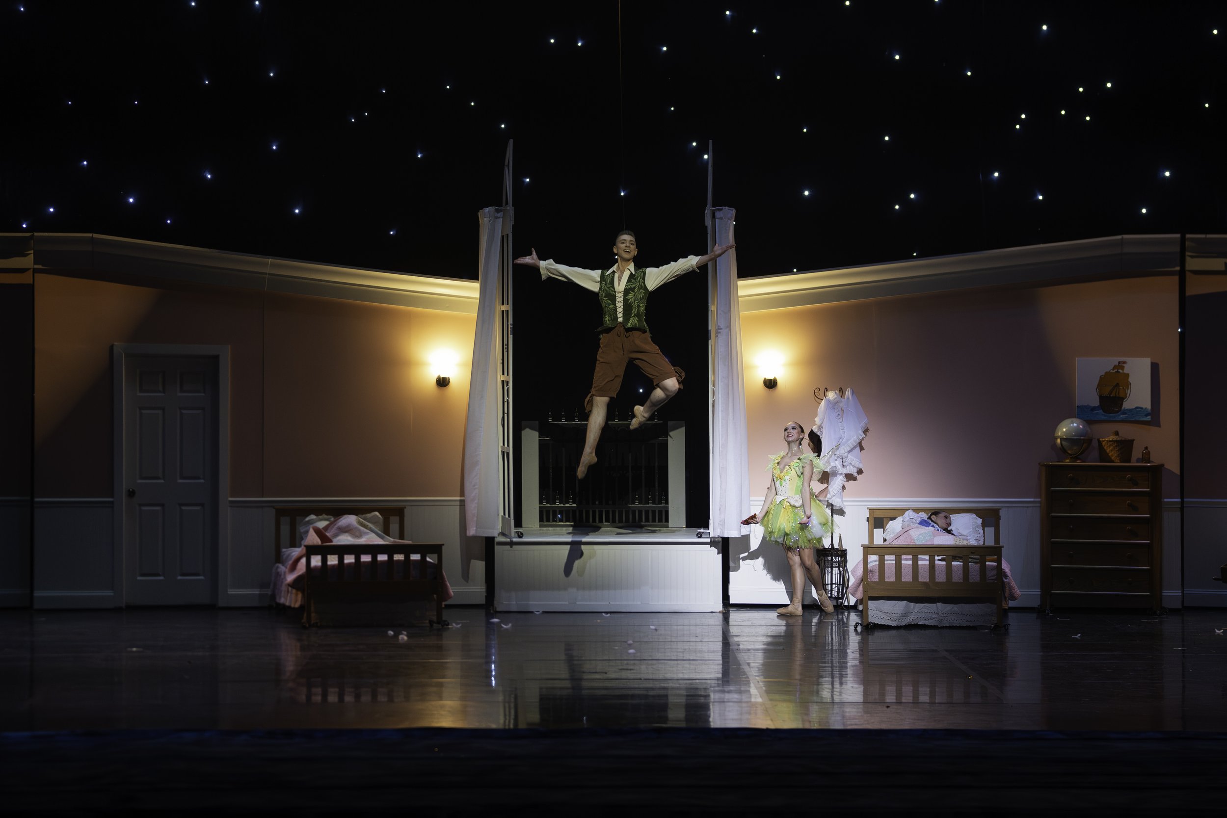 A stage scene from a theatrical performance set in a bedroom with pink walls, including beds, a dresser, and wall decor. A male performer is mid-air, hanging from a trapeze, with arms outstretched. Two female performers are on stage: one dressed in a