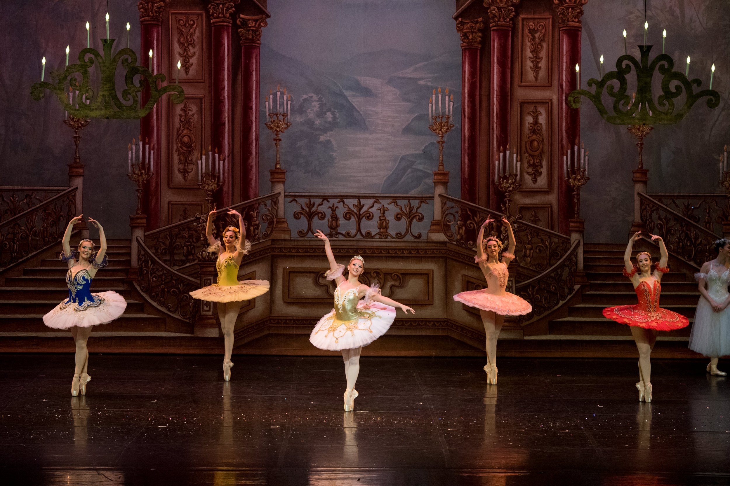 Ballet dancers performing on stage in colorful tutus in front of a grand, ornate set with staircase and chandeliers.