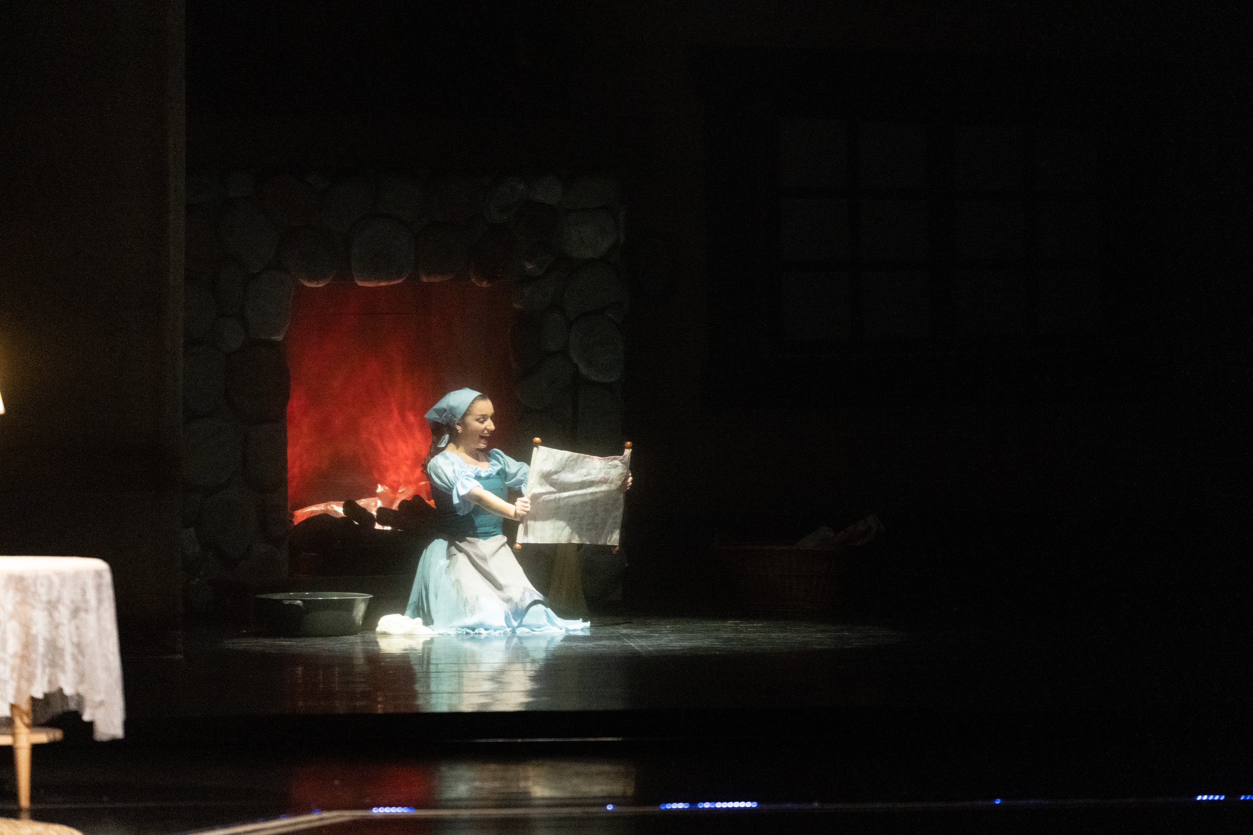 A woman dressed in historical clothing kneels beside a fireplace, holding a sheet of paper, with a warm orange glow illuminating her face.