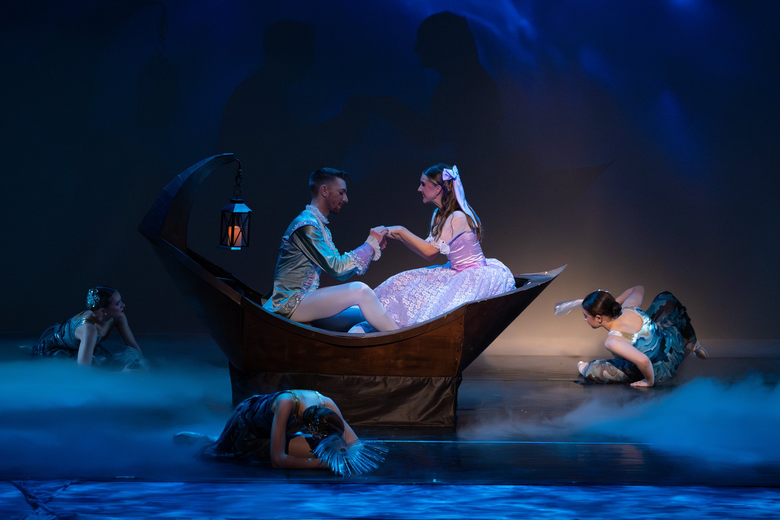 A theatrical stage scene with a man and woman in a boat, holding hands, surrounded by four dancers in costumes, with a foggy background and blue lighting.