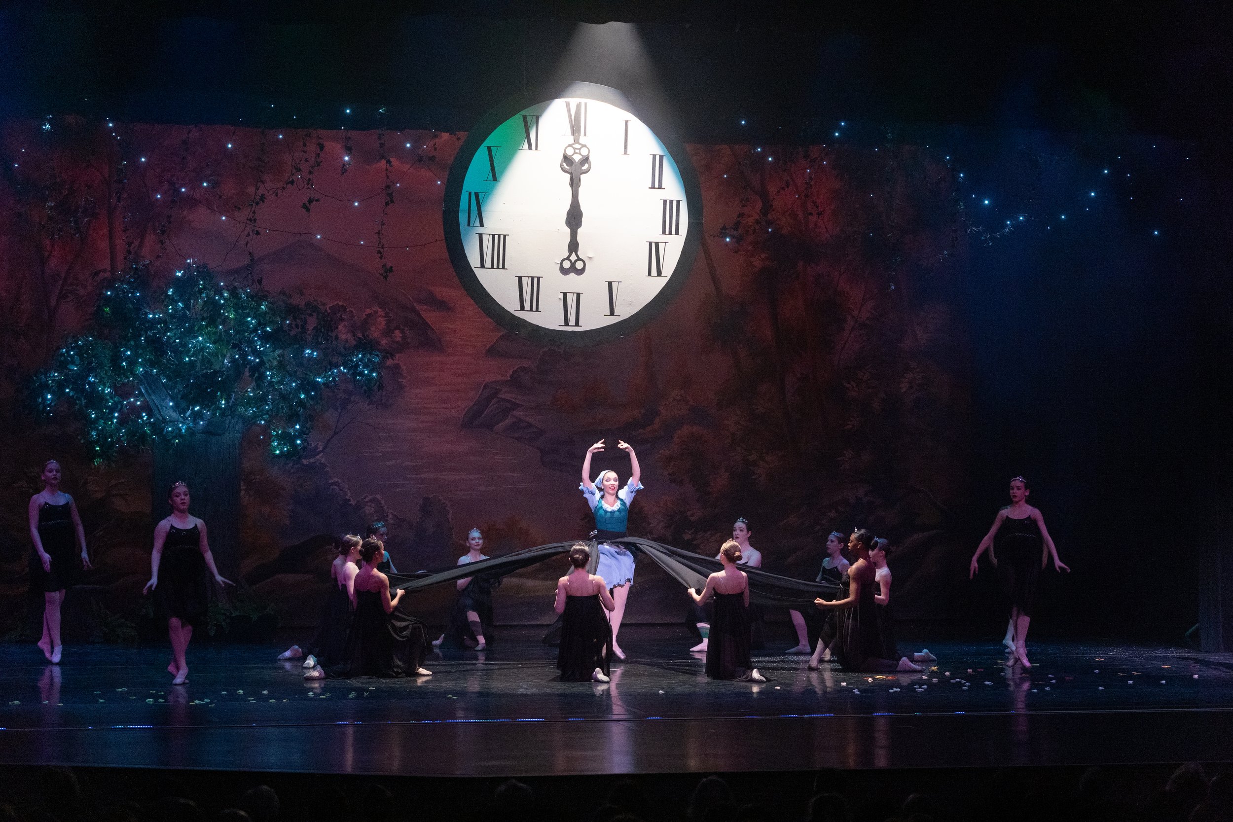 A ballet performance on stage with a large clock in the background showing midnight, surrounded by a painted scenic backdrop and illuminated tree. Dancers in black costumes perform, with one in a blue dress at center gesturing upward.