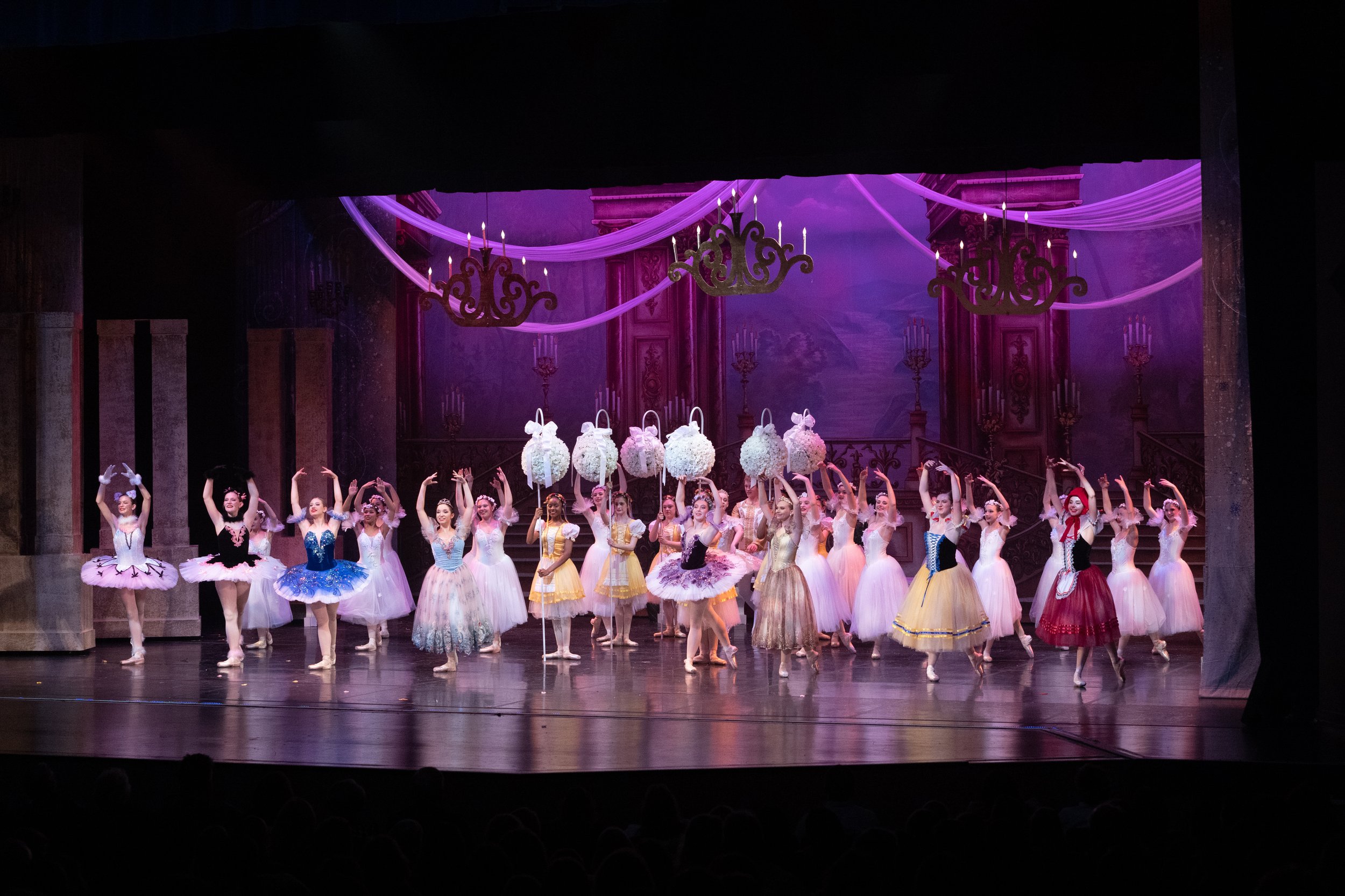 Ballet dancers in colorful tutus performing on a stage with ornate purple and gold backdrop and chandeliers.