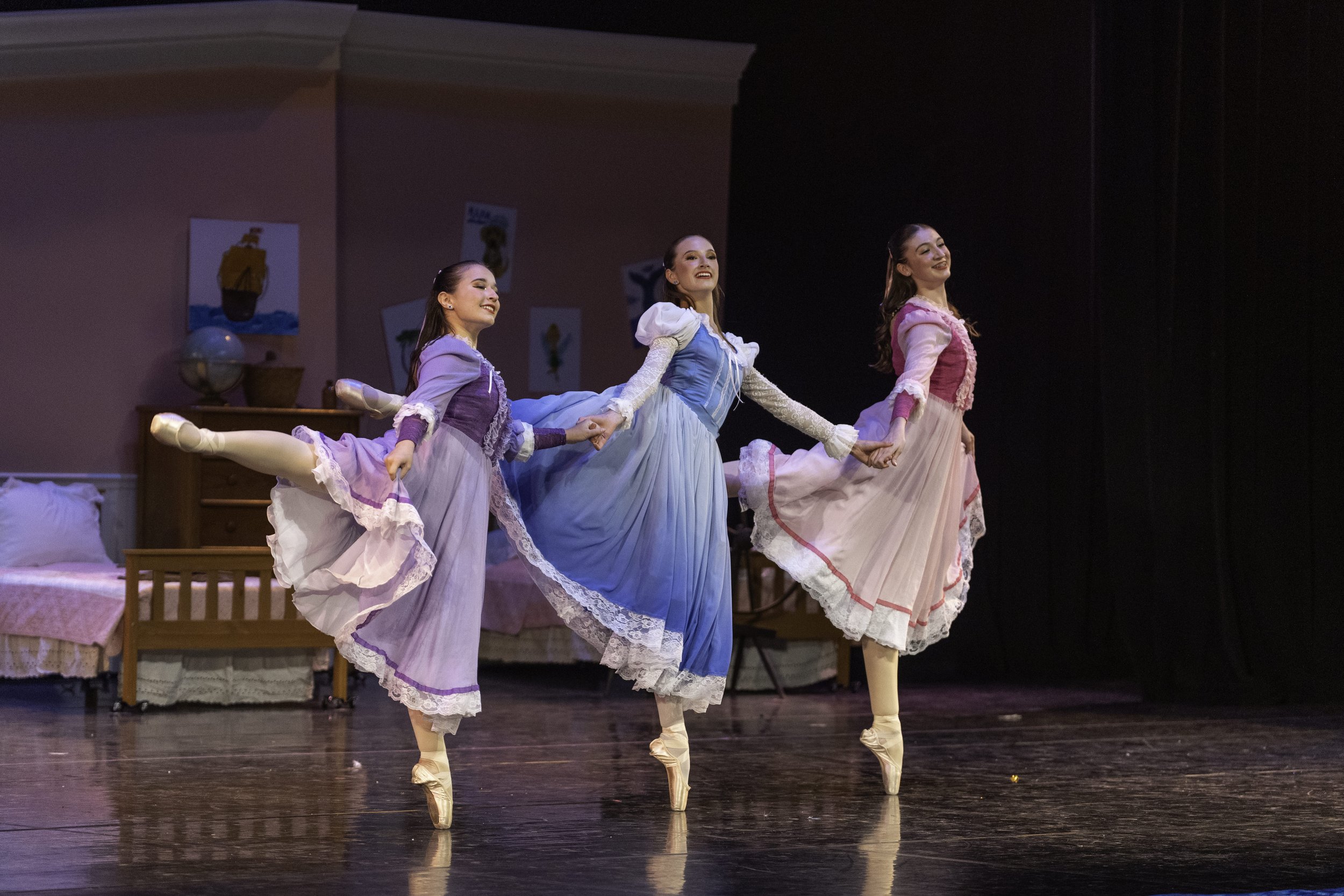 Three ballerinas in pastel-colored dresses performing a dance on stage, holding hands and balancing on tiptoes, with a bedroom set and colorful artwork in the background.