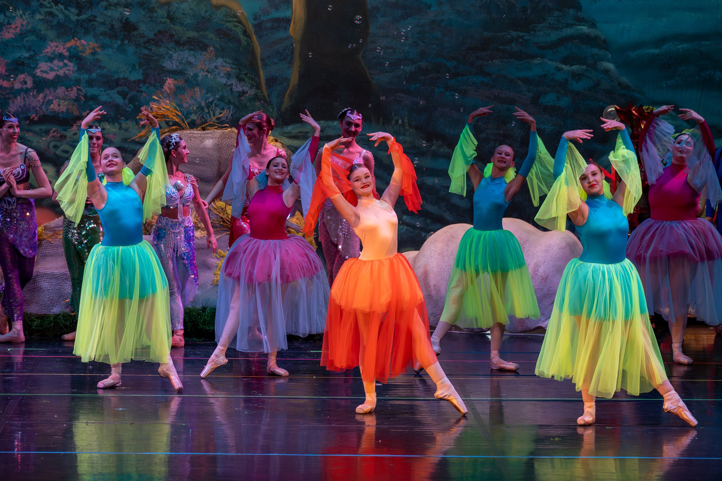 Ballet dancers in colorful tutus performing on stage with a nature-themed backdrop.