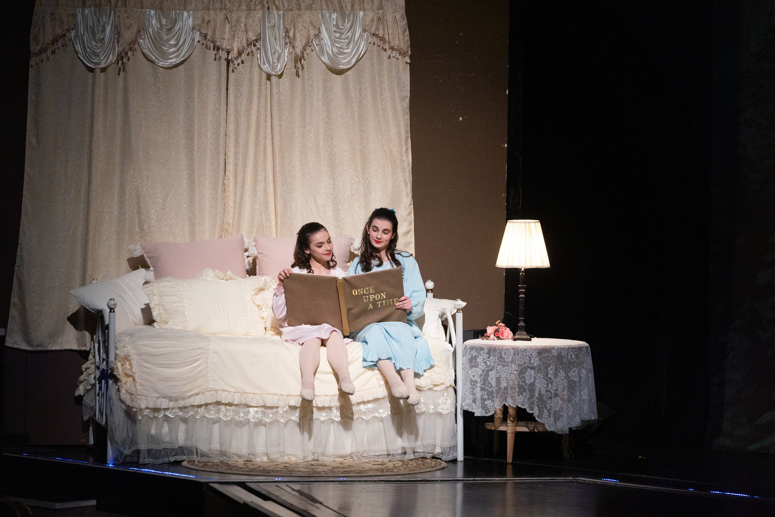 Two young women dressed as princesses sitting on a bed, reading a storybook titled 'Once Upon a Time', in a decorated bedroom setting with vintage curtains, pillows, a lamp, and a small table with flowers.