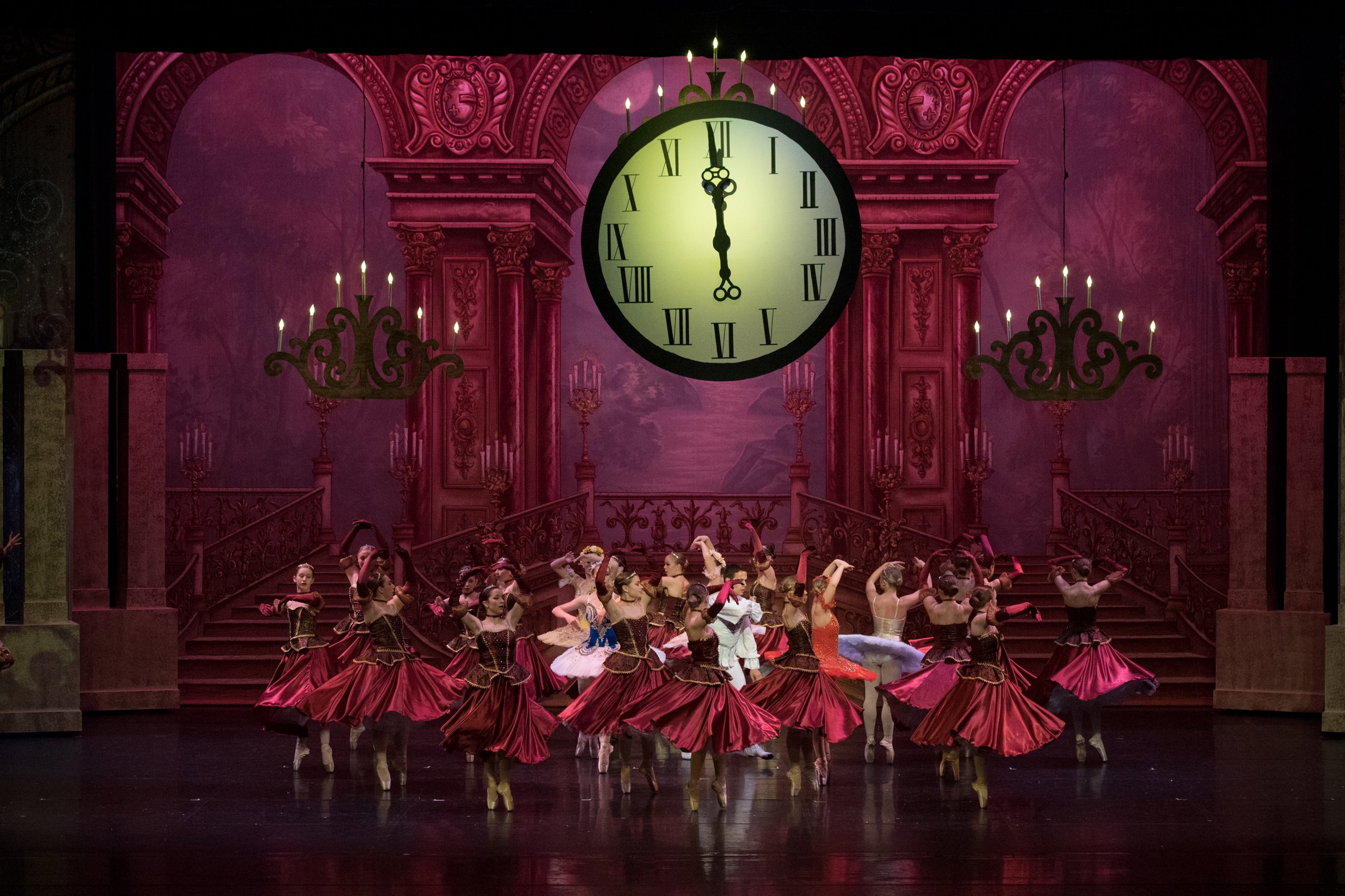 A ballet stage depicting a scene from 'Cinderella' with dancers in burgundy and white costumes, a large clock striking midnight in the background, and ornate chandeliers and staircase set design.