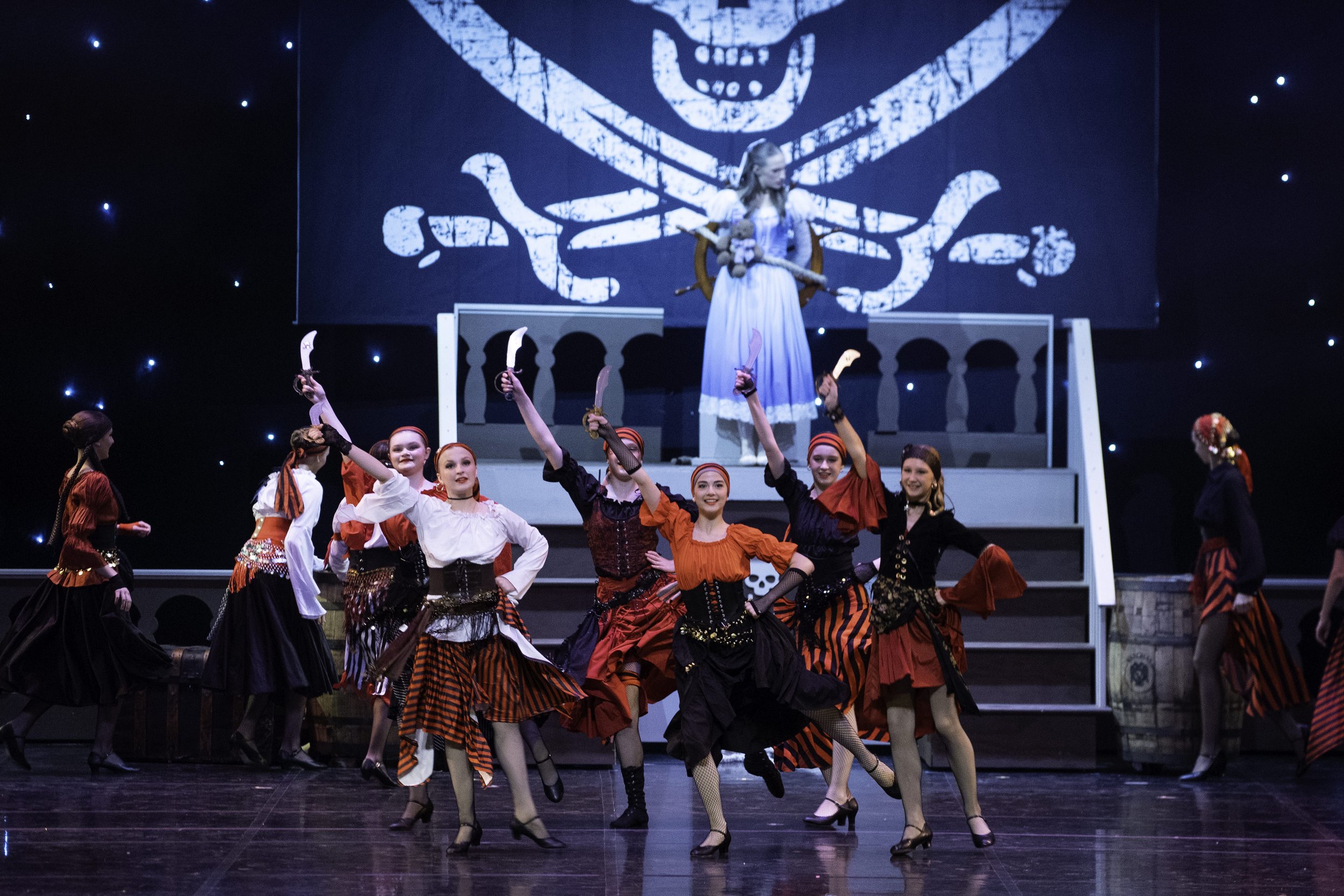 Stage scene with women in pirate costumes performing a dance. A woman dressed as a pirate with a sword is on a raised platform in the background, with a large pirate flag projected behind her. The performers on stage are in red, black, and white cost
