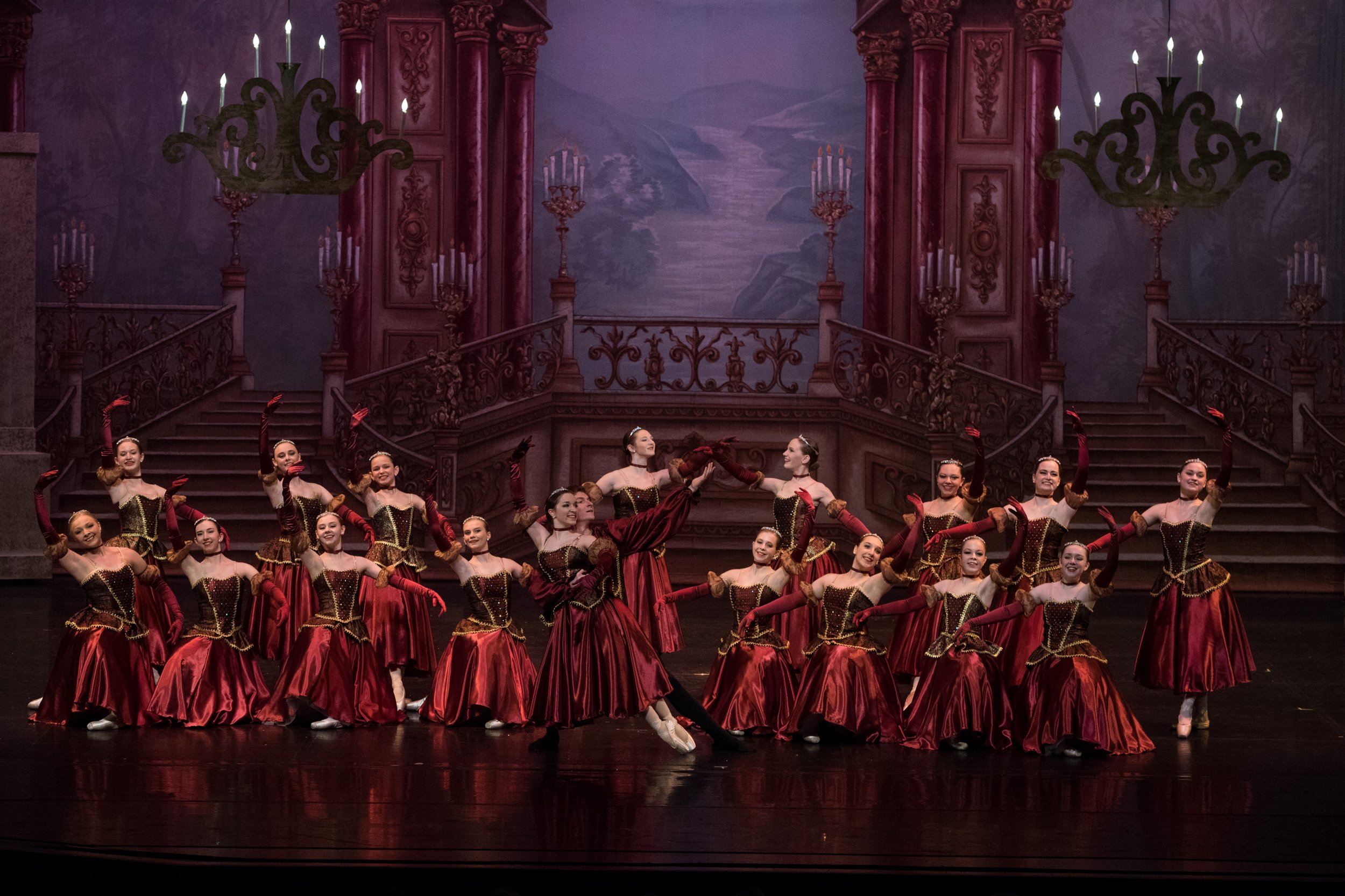 A ballet performance with dancers in red and gold costumes on a stage with a elaborate castle backdrop and chandeliers.
