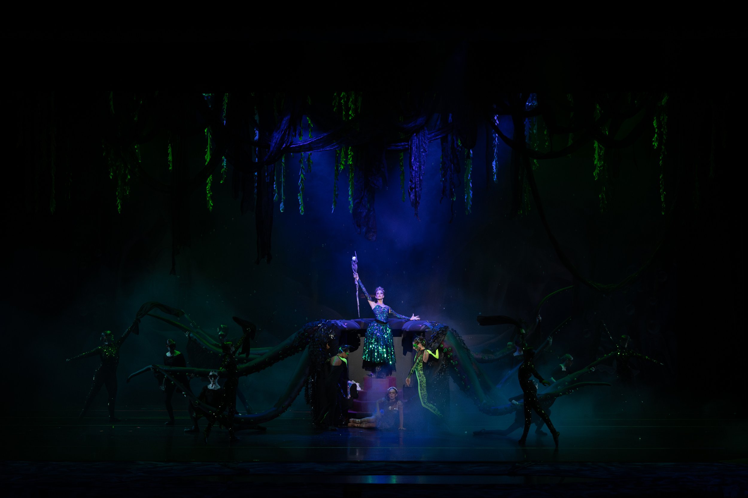 A theatrical stage performance with dancers in costume, set against a dark background with hanging fabric-like decorations and lighting highlighting the central performer.