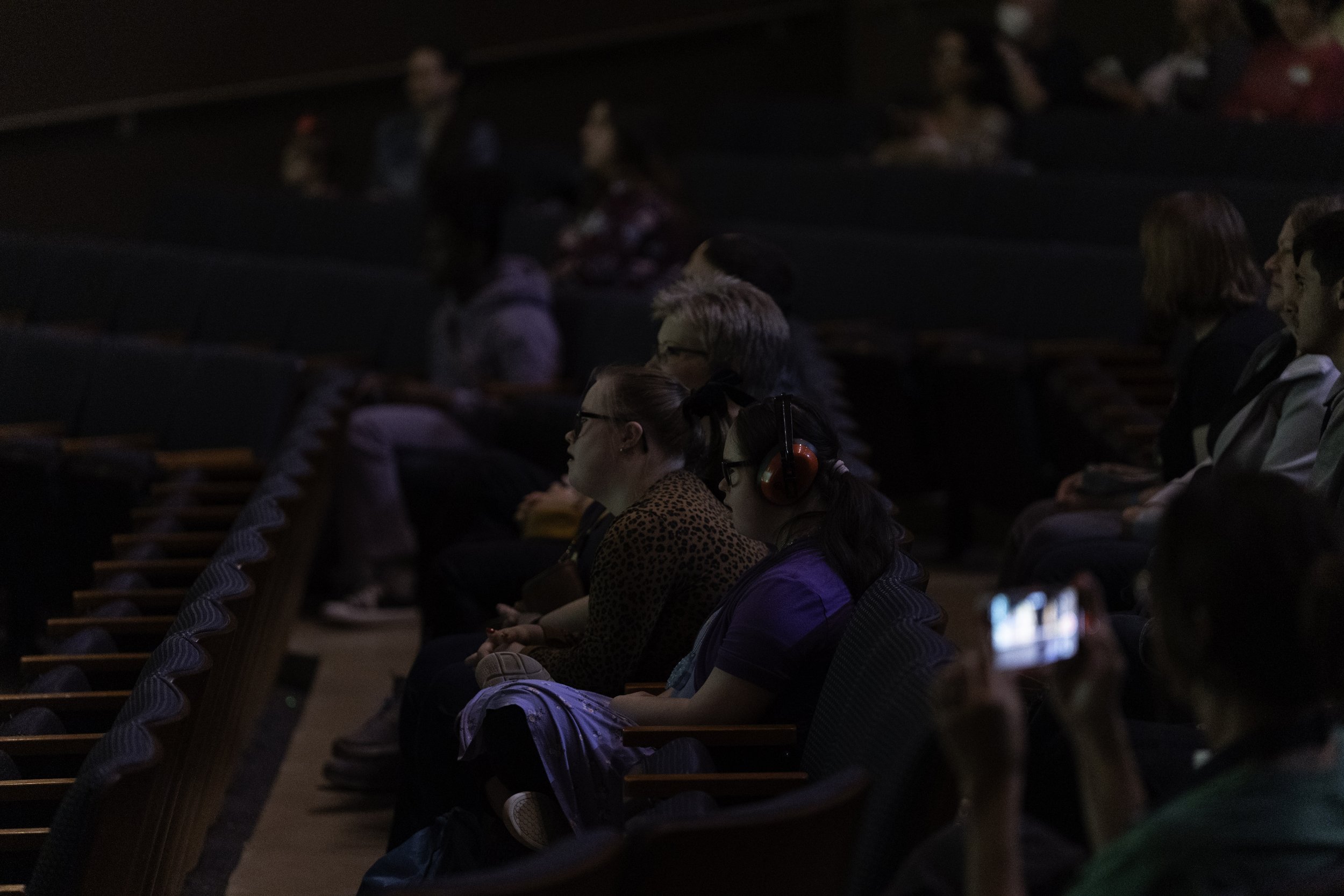 Audience members sitting in a darkened auditorium, some wearing headphones, watching a presentation or performance.