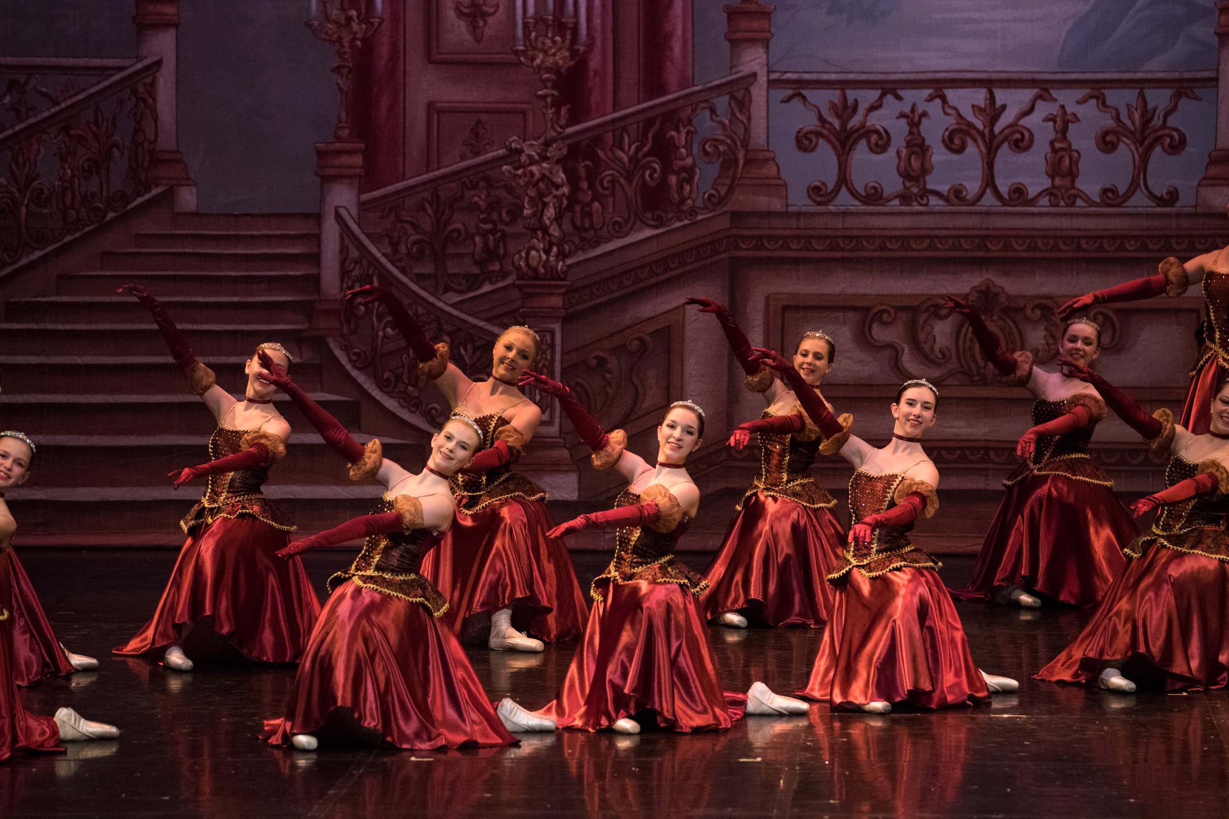 A group of female ballet dancers in red and gold costumes performing on stage with a painted palace staircase backdrop.