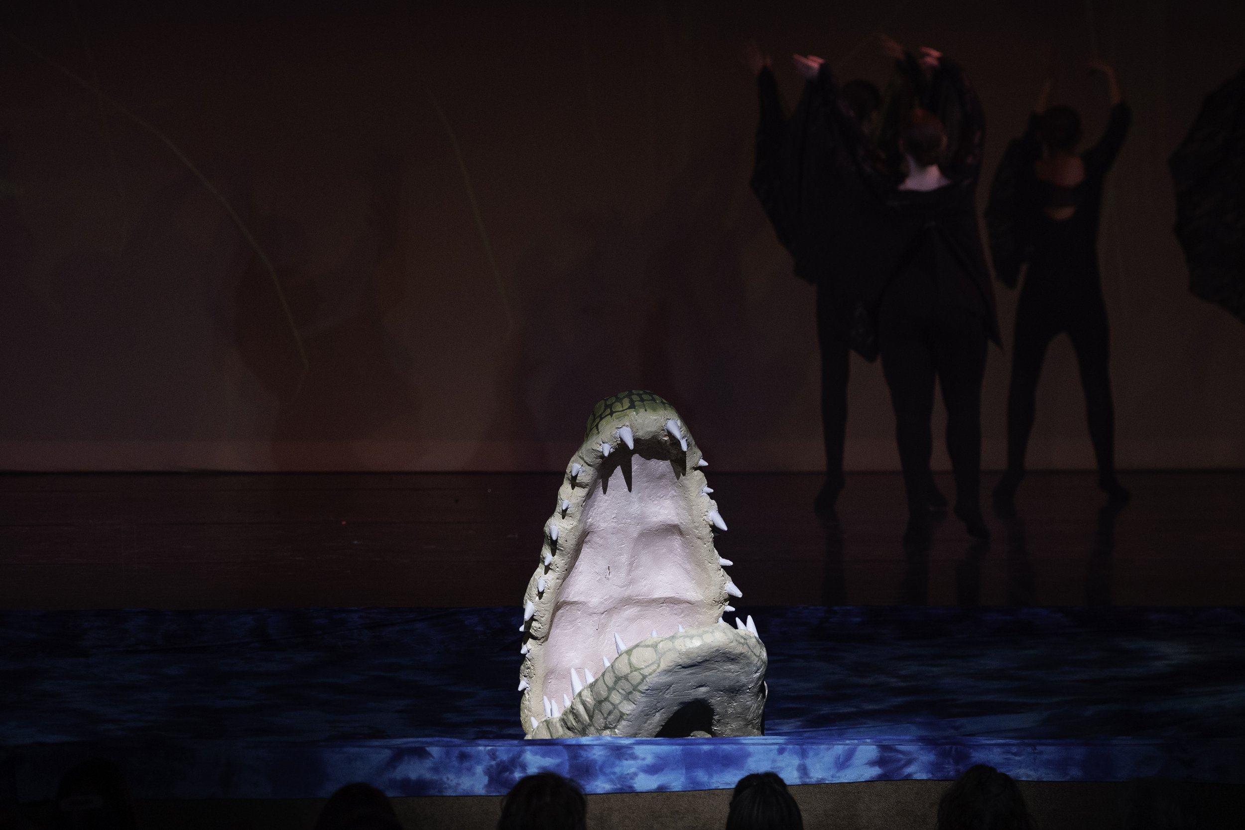 A stage with a crocodile prop with its mouth open, appearing to emerge from water, with dancers in black costumes in the background.