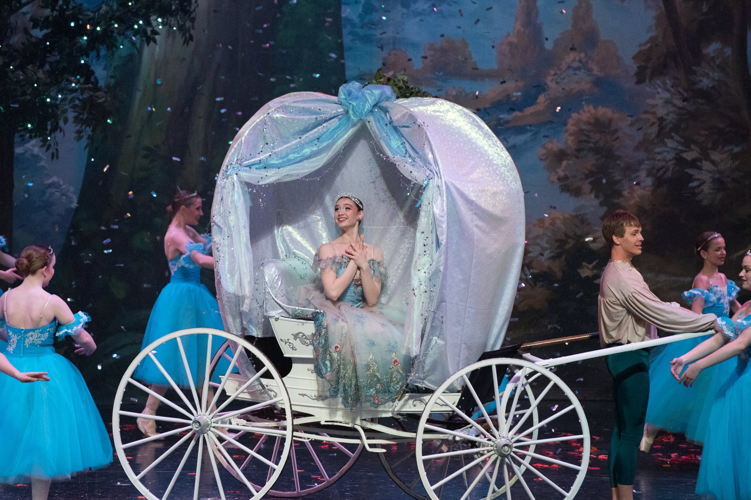 A ballerina dressed as Cinderella in a silver and blue costume sitting in a white fairy tale carriage on stage, surrounded by dancers in blue tutus and a man in beige attire, with a scenic backdrop and confetti.