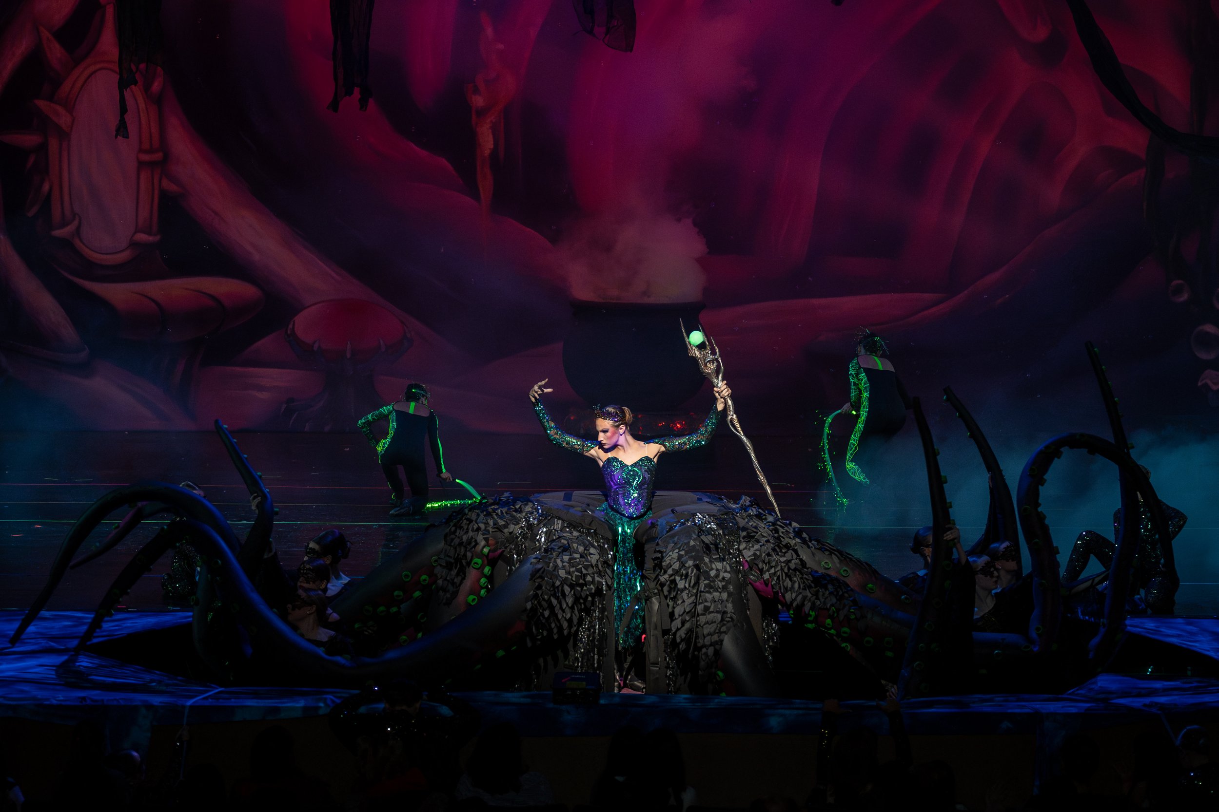 A theatrical stage performance featuring a performer dressed as a mermaid with a large, elaborate costume resembling an octopus, holding a staff, surrounded by other performers with neon accents, set against a dark, colorful, fantastical backdrop wit