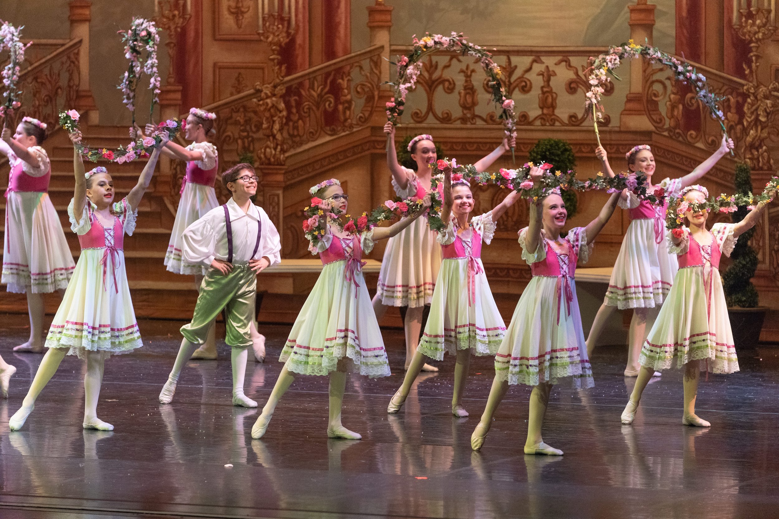 Children performing a ballet on stage, dressed in traditional costumes with floral accents, under a decorative wooden backdrop.