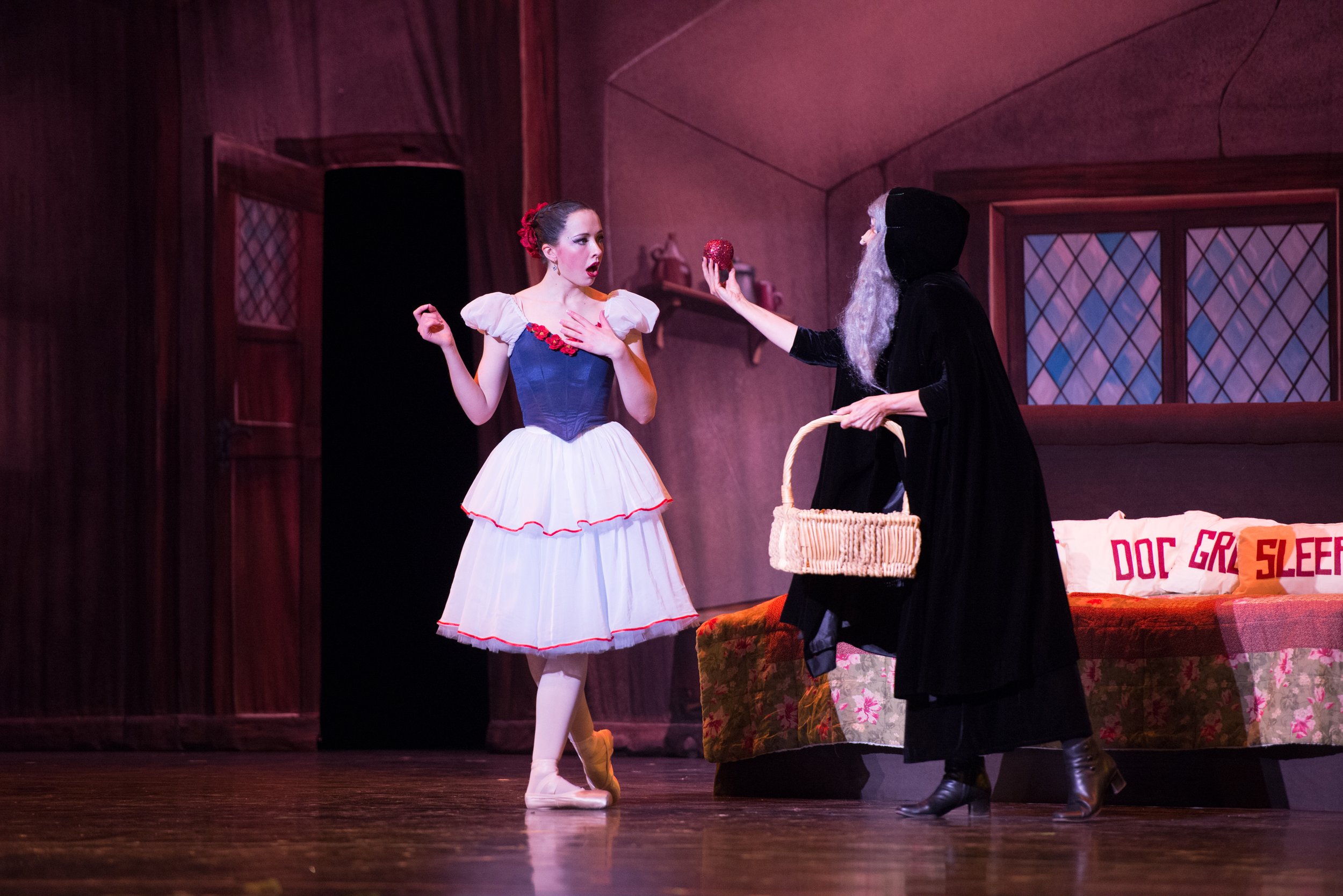 A theatrical performance with a dancer dressed as Snow White and an actor dressed as a witch on stage. Snow White looks surprised, touching her chest, while the witch holds out a red apple and carries a basket. The backdrop shows a bedroom set with a