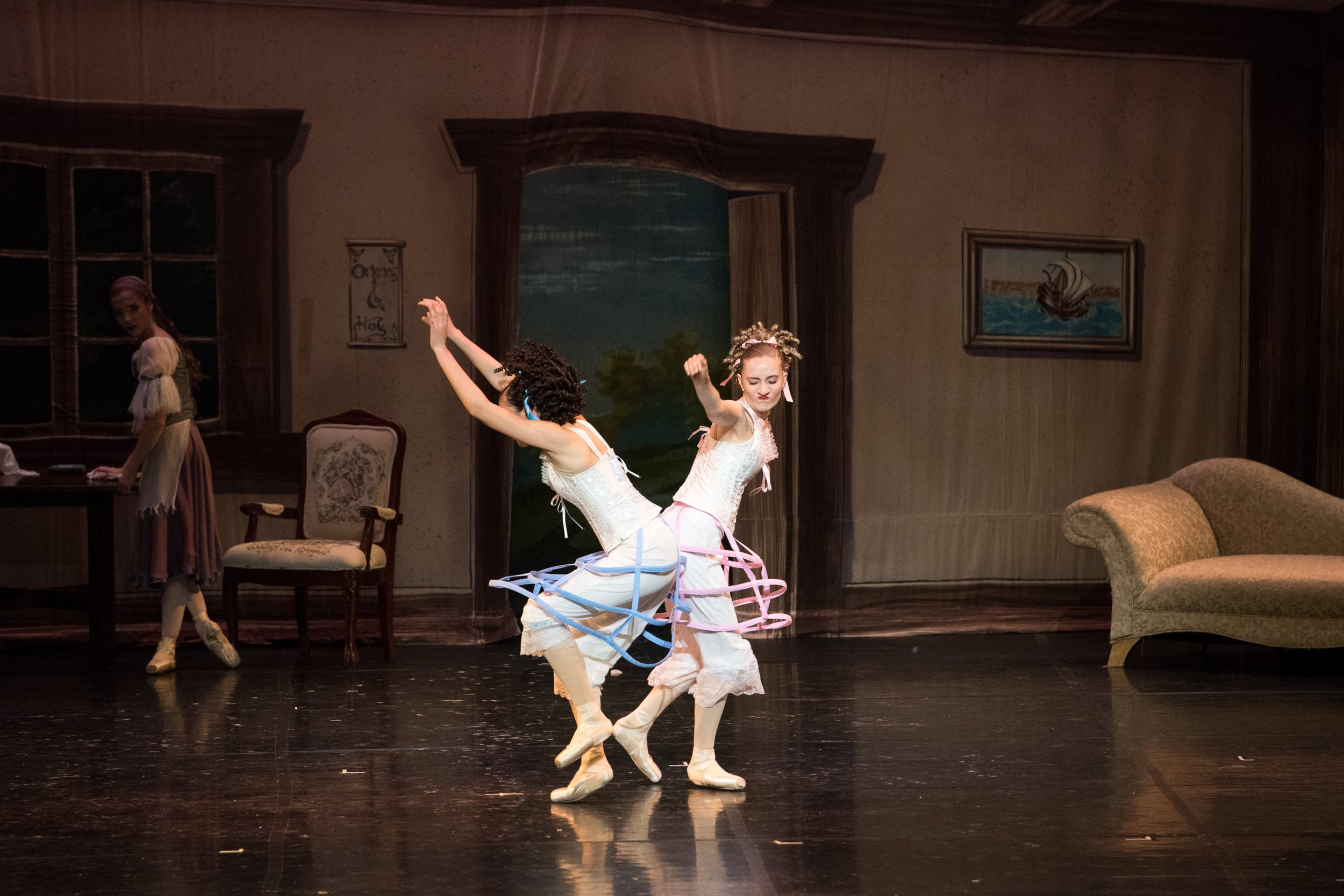 Two ballerinas with colorful hoop skirts performing on stage in front of a painted backdrop, with a third person observing in the background.