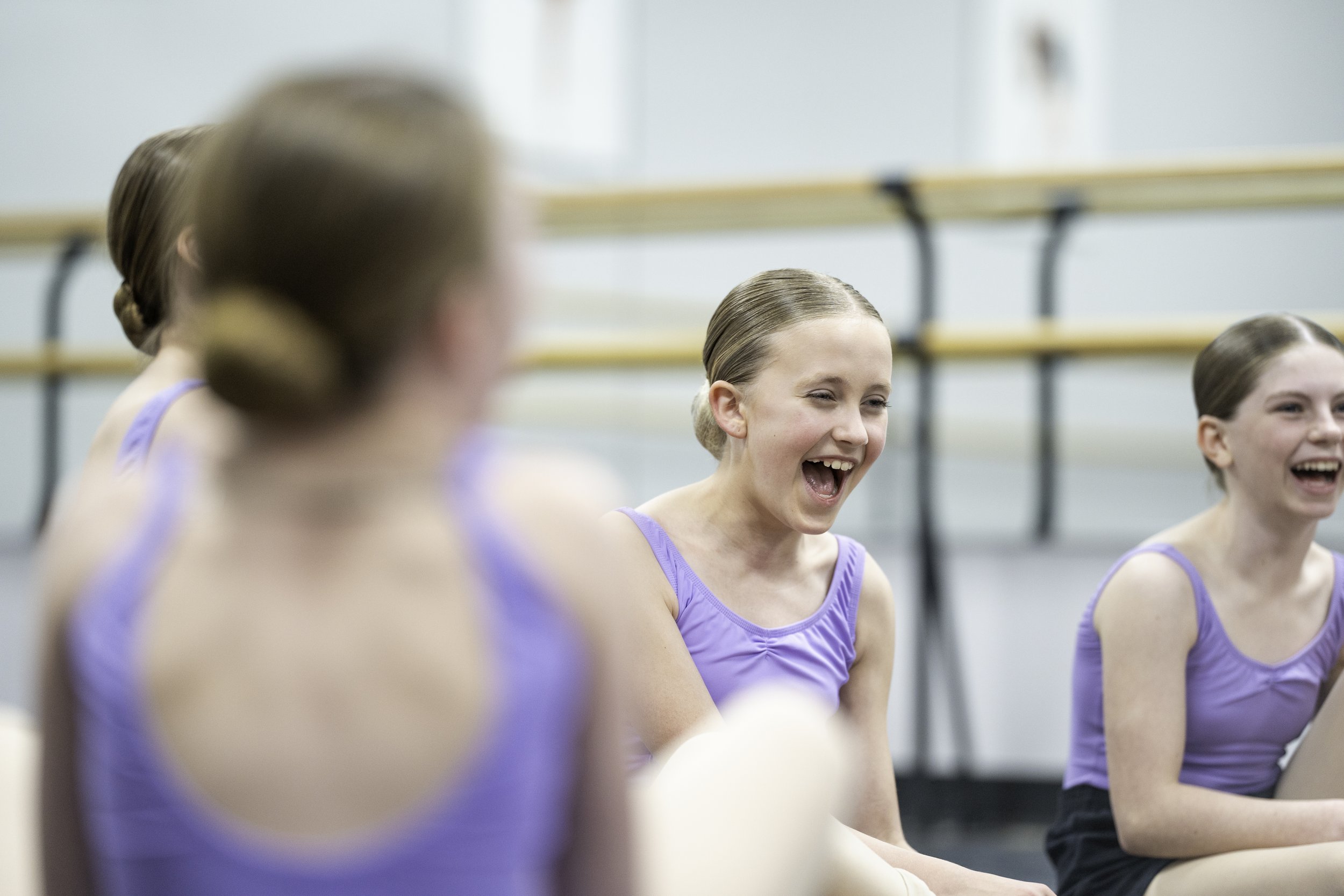 Group of young girls in purple dance costumes sitting and laughing during dance class or rehearsal.