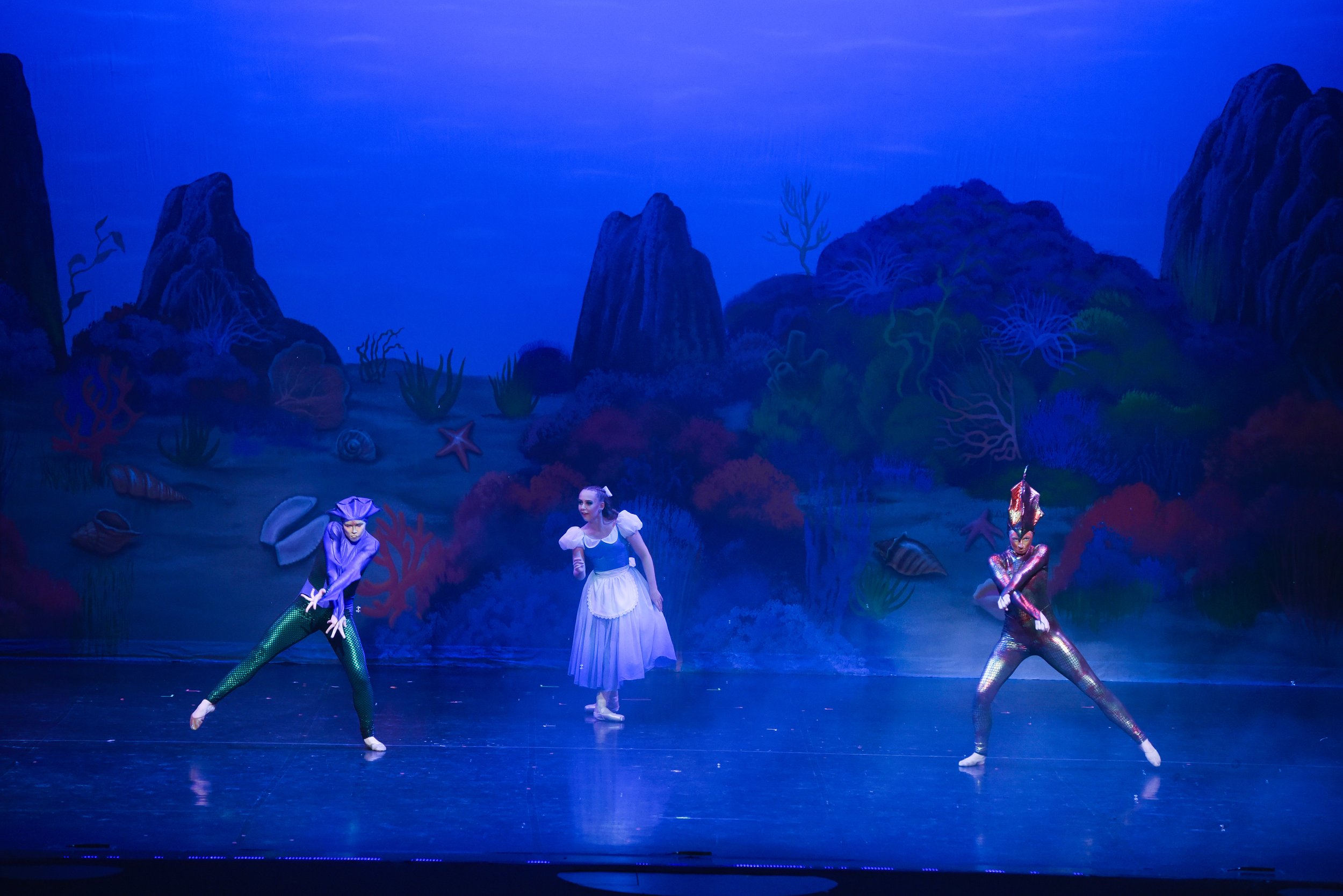 Three dancers performing on a stage with an underwater themed backdrop, featuring coral, shells, starfish, and rocks, under blue lighting.