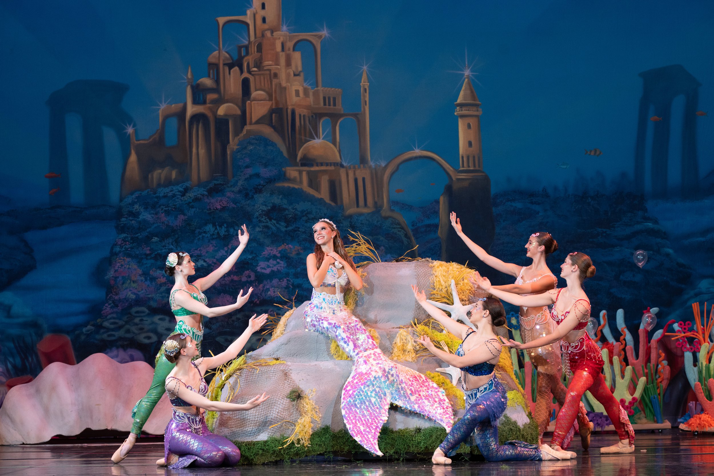 A theatrical dance performance featuring young women dressed as mermaids on stage with an underwater castle backdrop.