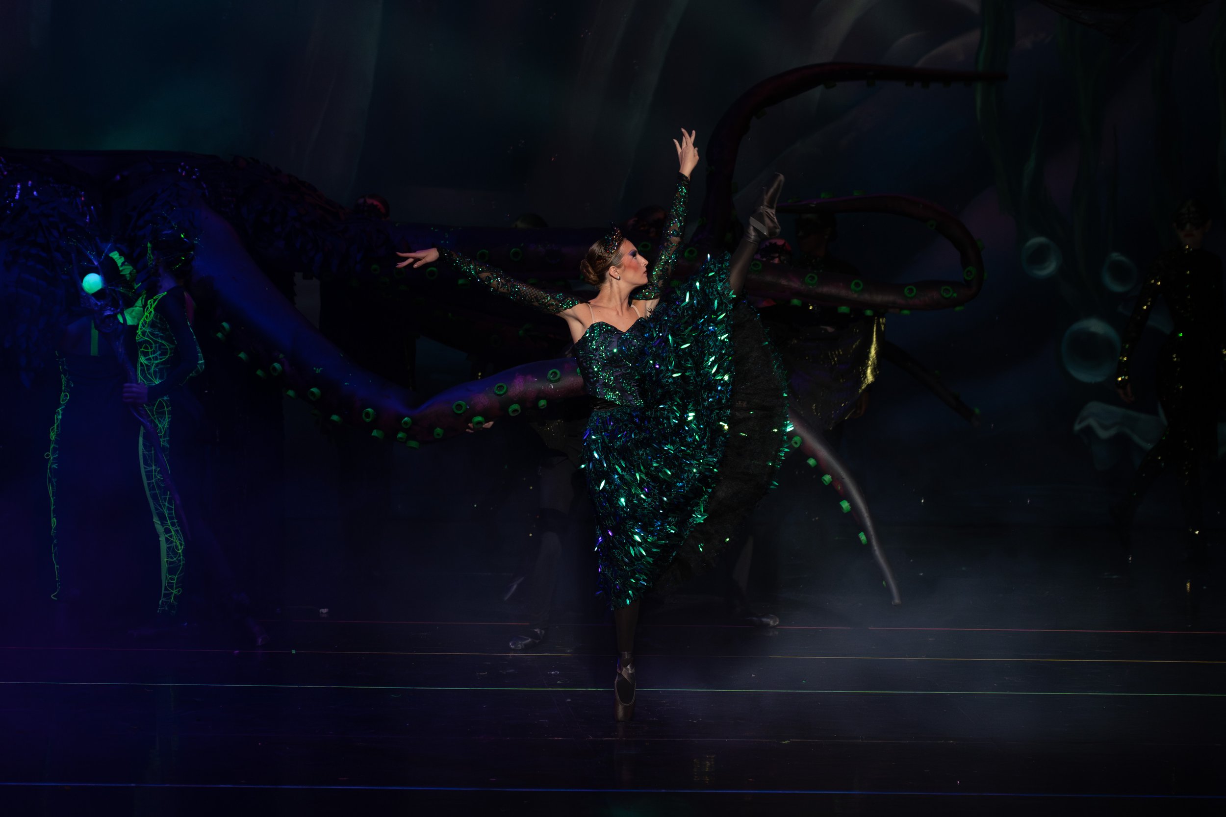 A performer in a glittering green and black costume dances on stage with a large, twisted, tentacle-like prop in the background, illuminated by dramatic stage lighting.