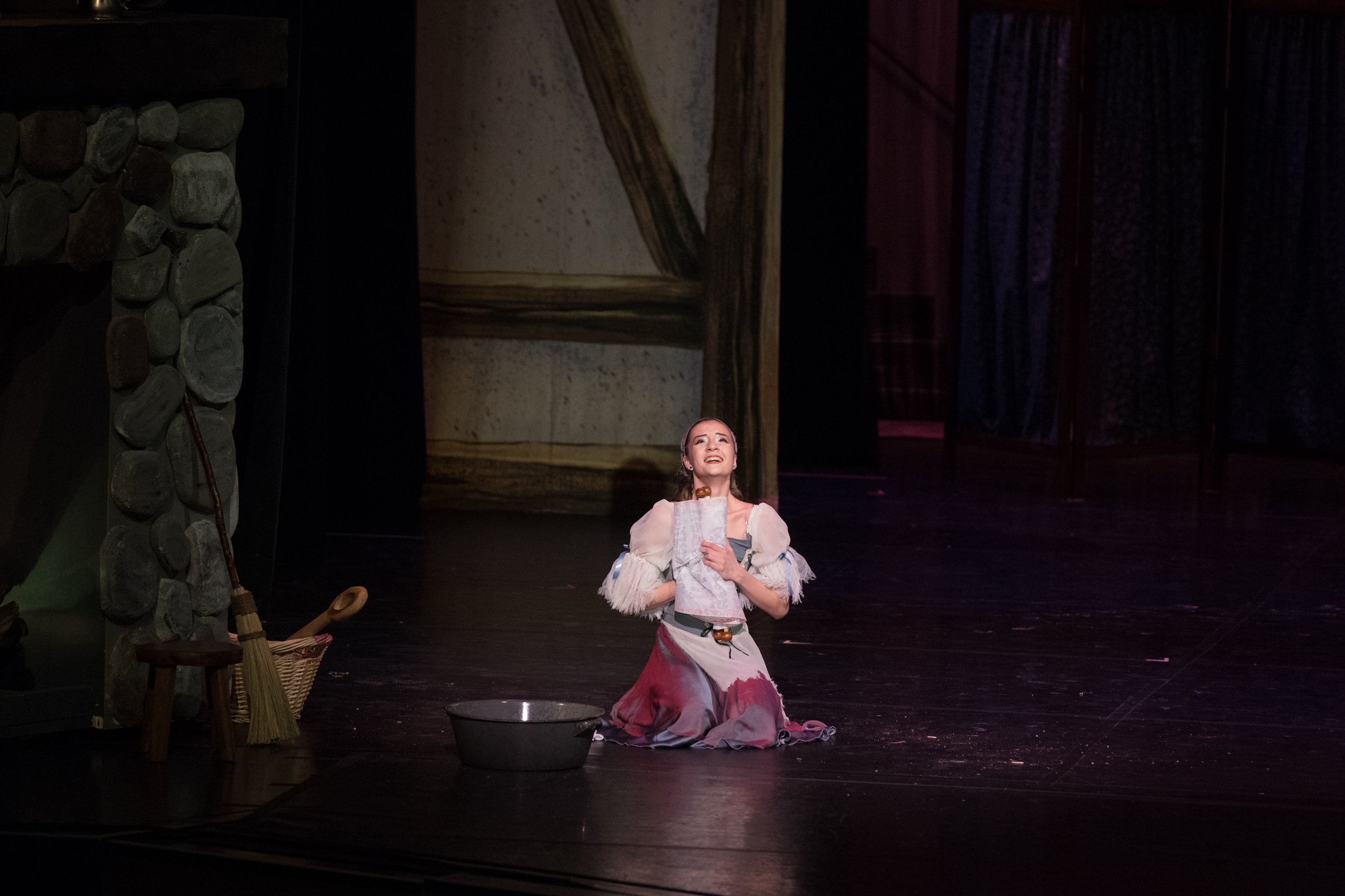 A woman dressed in costume kneels on stage, singing or speaking with a joyful expression, surrounded by stage props including a stone-looking wall, a small stool, a broom, a basket, and a basin, with a painted backdrop.