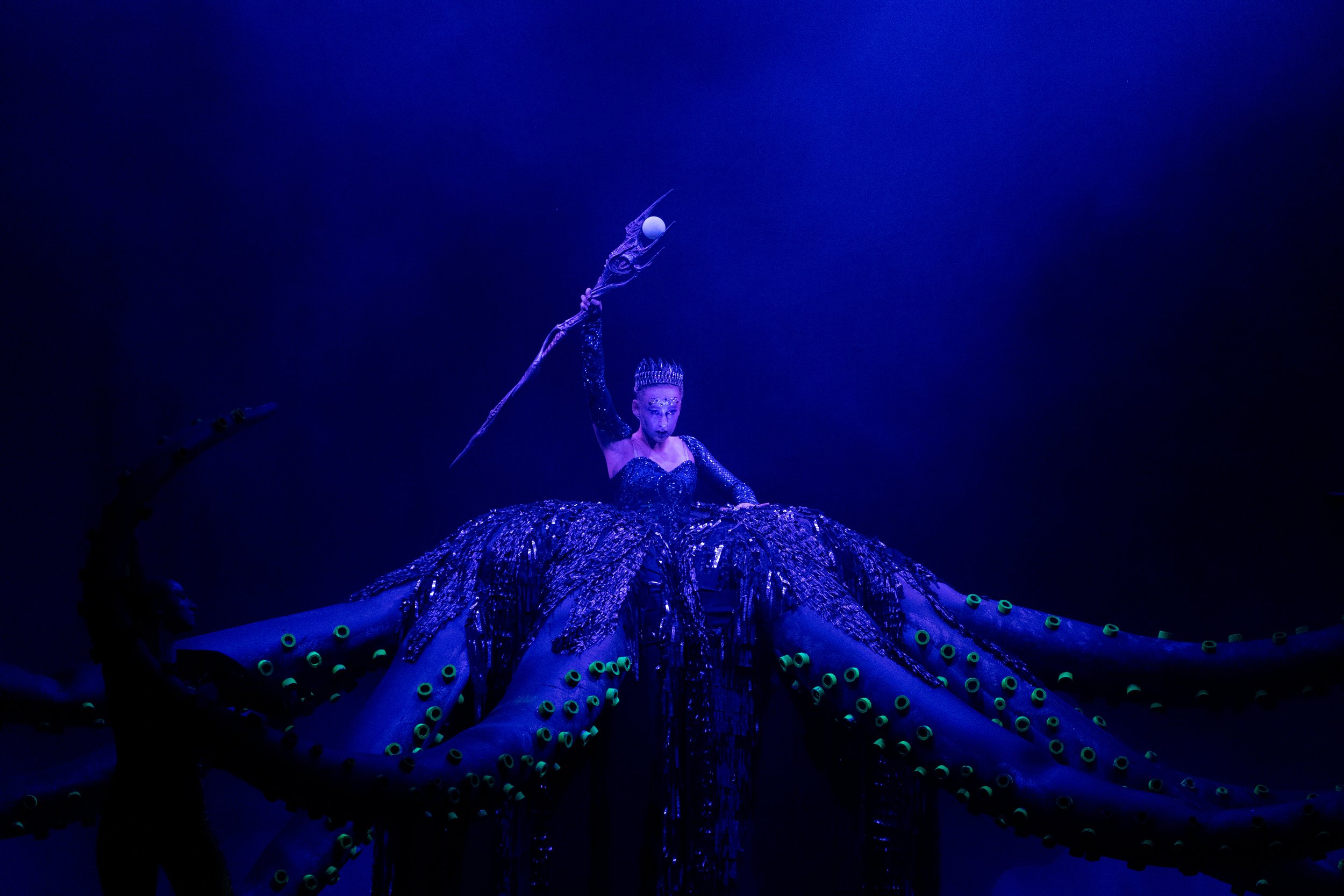 A performer in a dark, blue-lit setting wearing a black and blue costume resembling an octopus with large tentacles, holding a staff with a globe and a holographic projection, creating an artistic and theatrical scene.