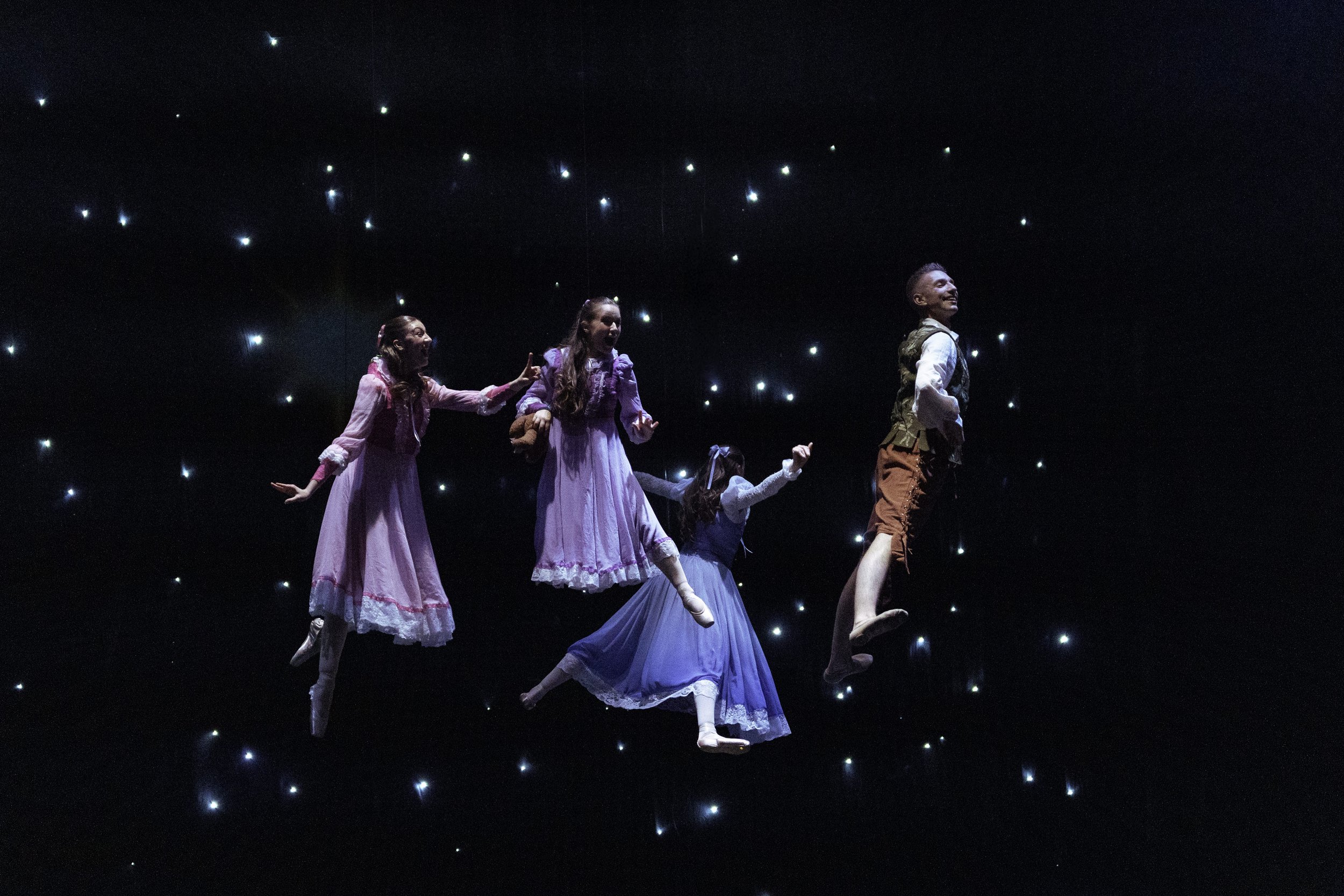 Four children in period costumes seemingly floating in a dark space with star-like lights, on stage during a theatrical performance.