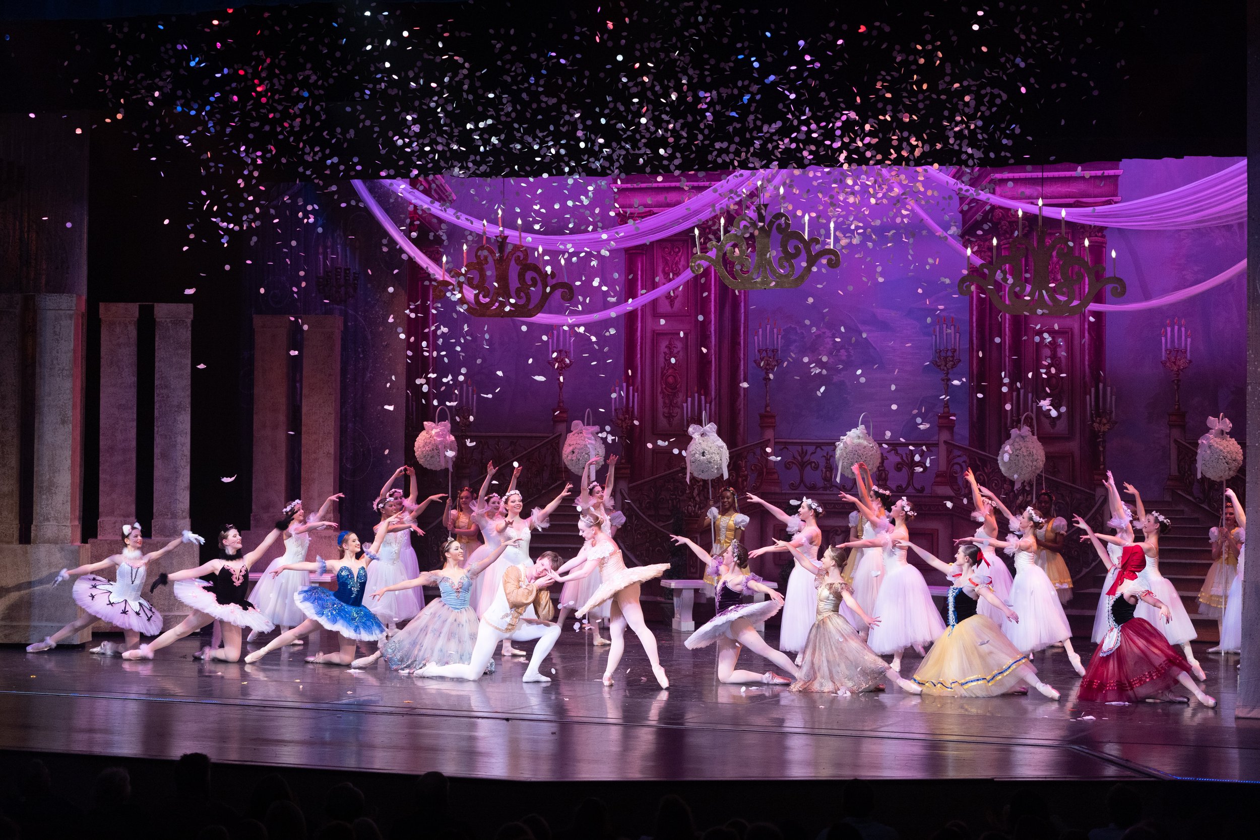 Ballet dancers in colorful costumes performing on stage with purple and pink decor and confetti falling.