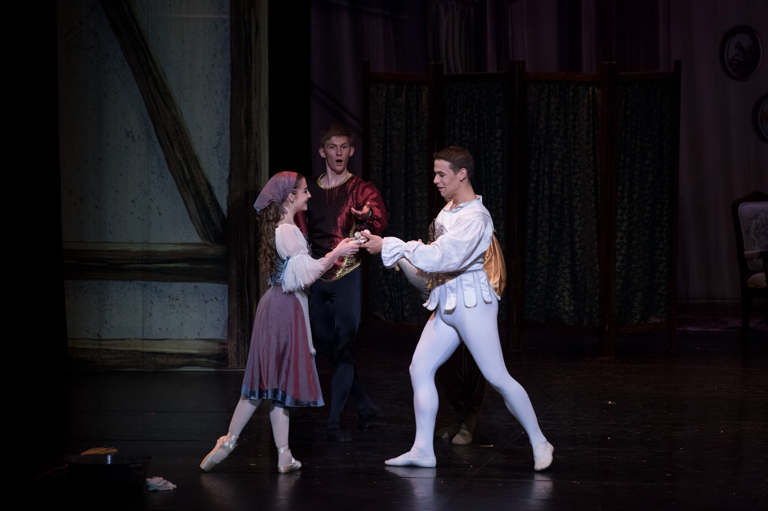 Three ballet dancers in period costumes performing on stage during a play or ballet, with a backdrop of trees and a stage set.