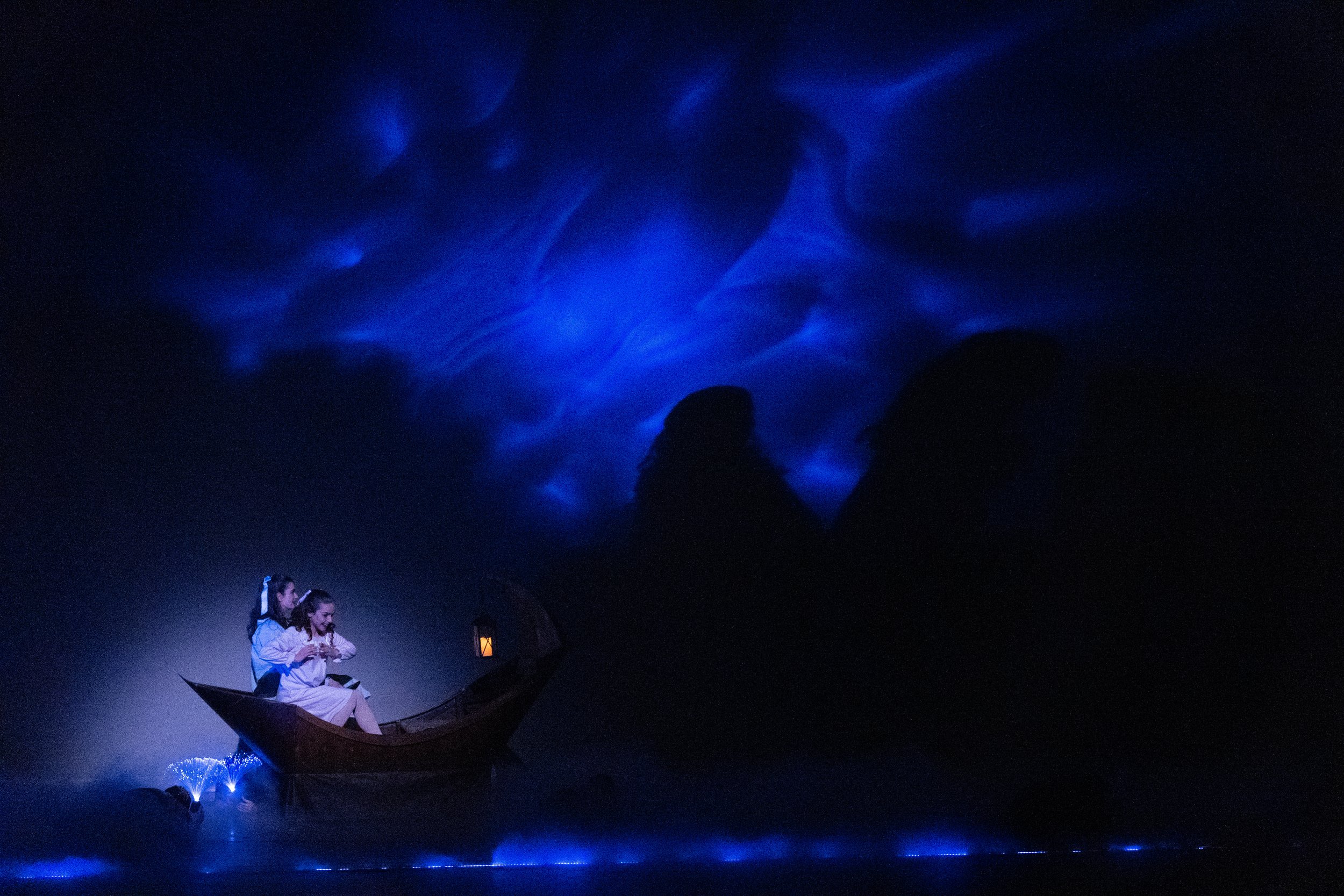 Two children sitting in a small wooden boat illuminated with a small lantern, surrounded by blue glowing water and sky effects, creating a magical night scene.