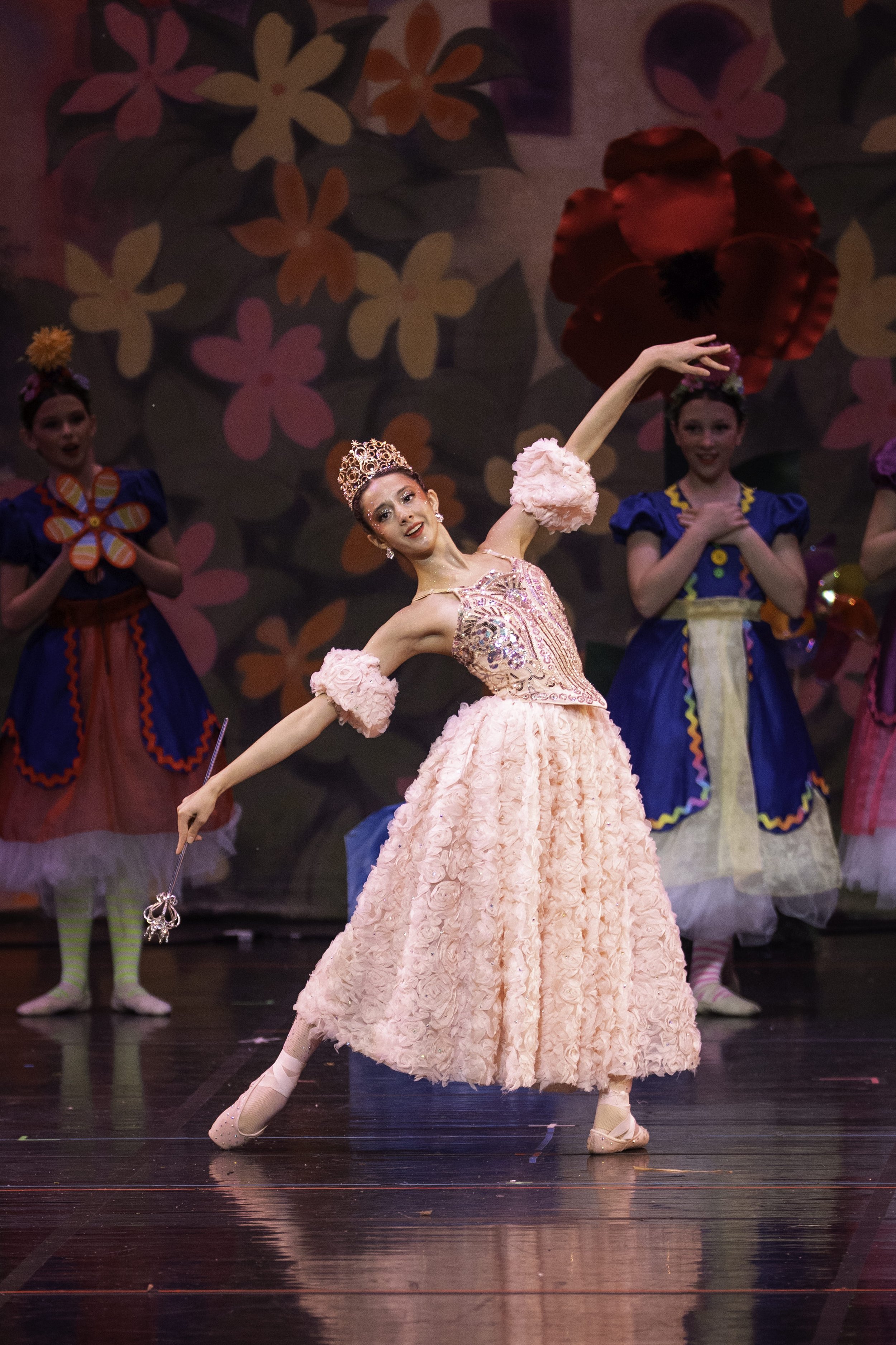 A ballerina in a pink, floral textured gown and a crown performs on stage, surrounded by young dancers in colorful costumes with flowers and butterflies in the background.