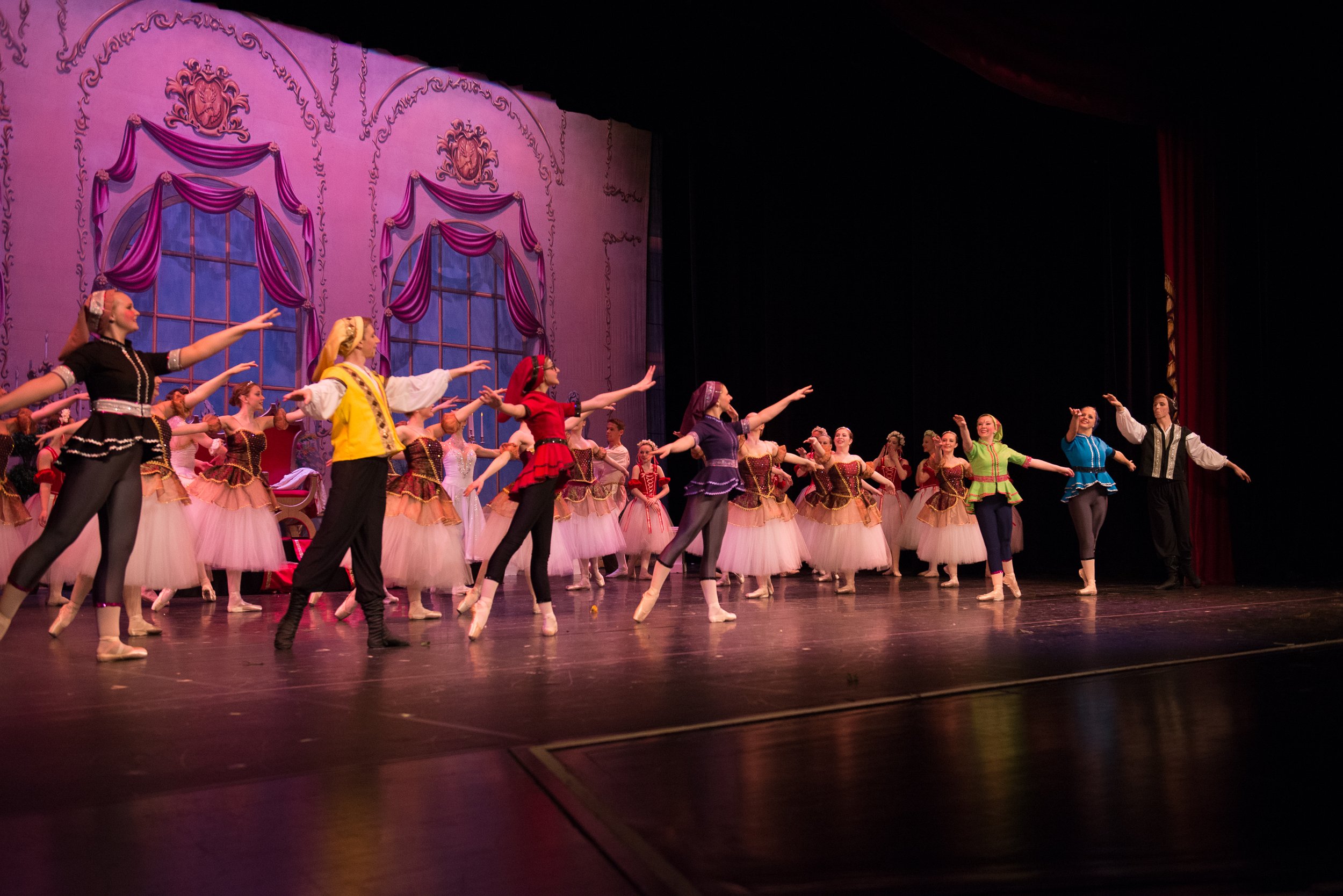 Ballet dancers perform on stage with pink and purple curtains and large windows in the background.