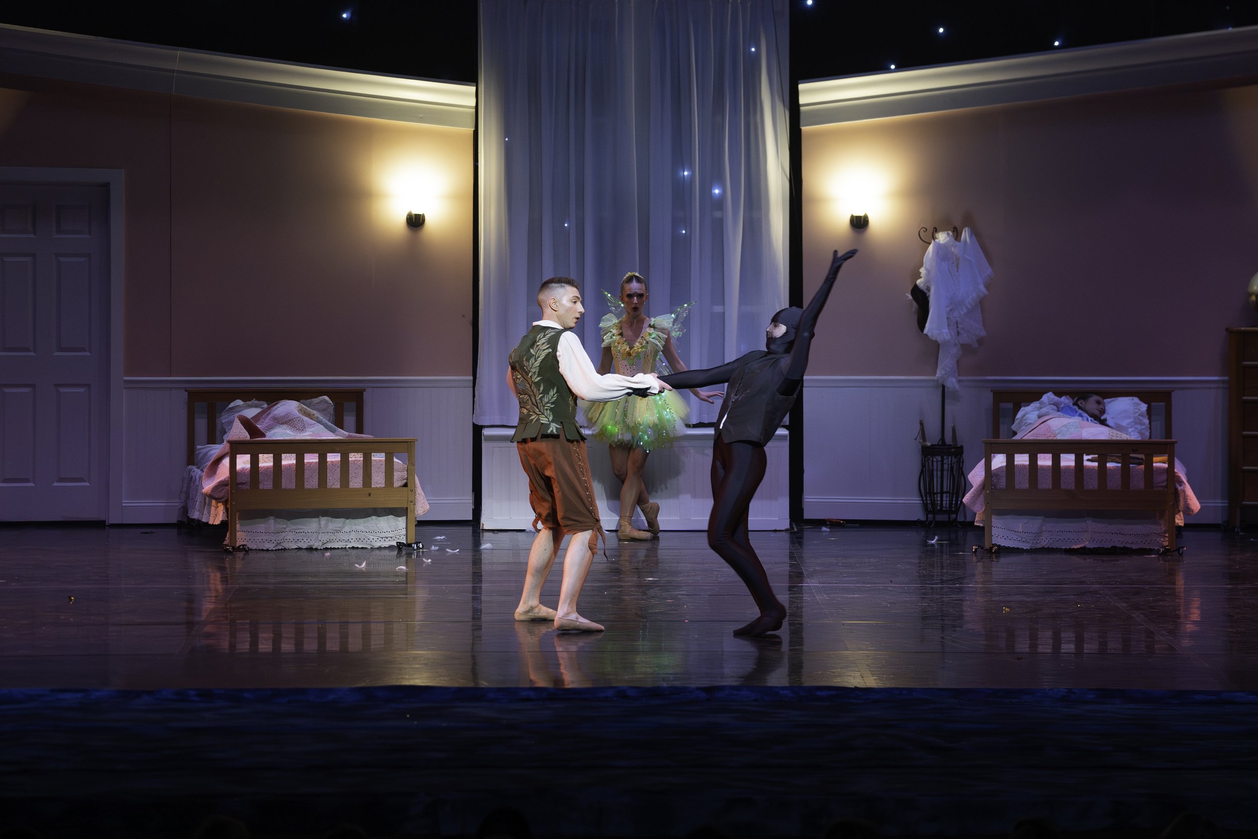 Group of ballet dancers performing on a stage set with beds and nightstands, with two beds and two actresses in costumes, two beds, one person in a fairy costume, and the other two in dance costumes dancing in the center of the stage.