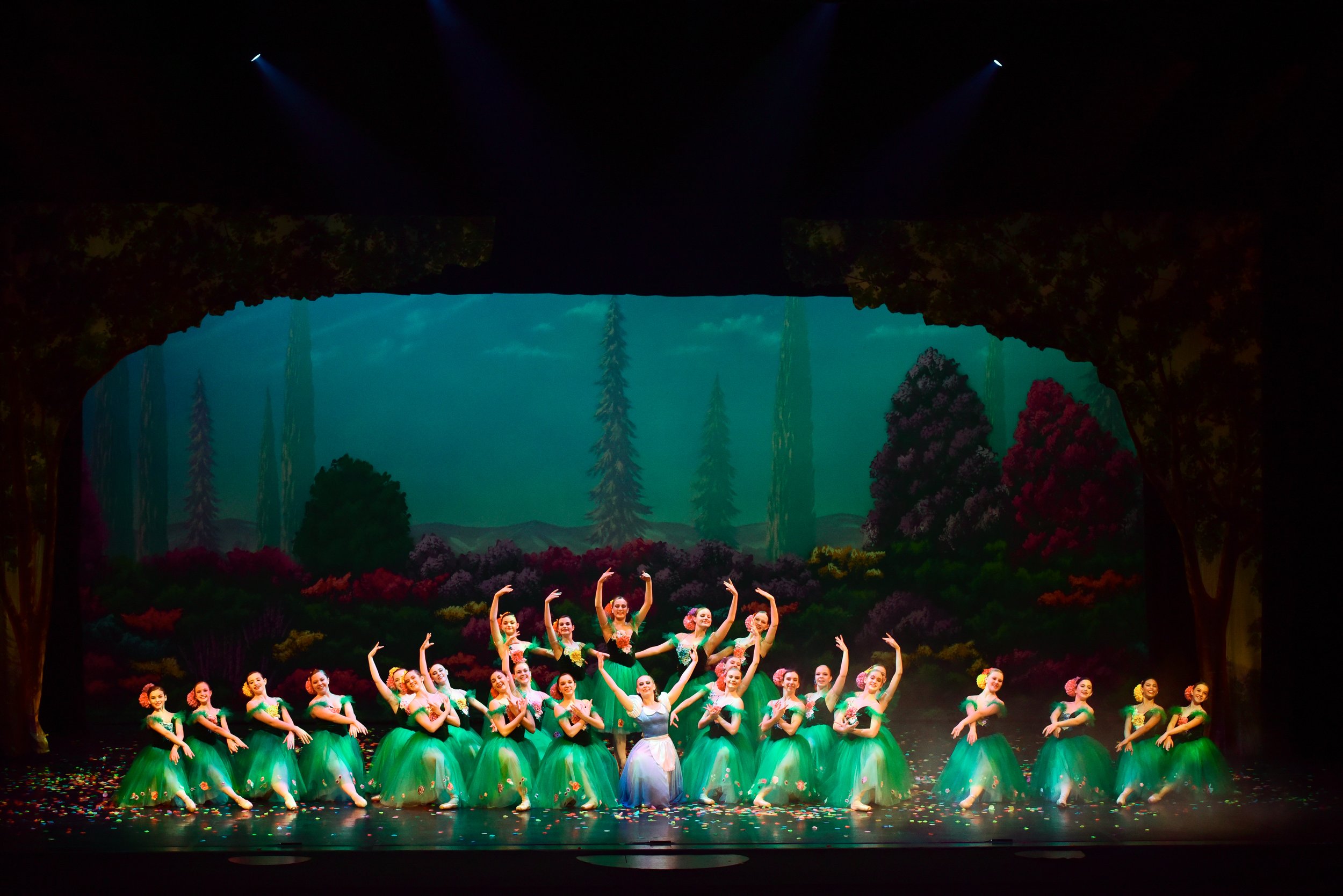 Ballet dancers on stage in green tutus performing in front of a scenic backdrop of trees and mountains, with confetti on the floor.