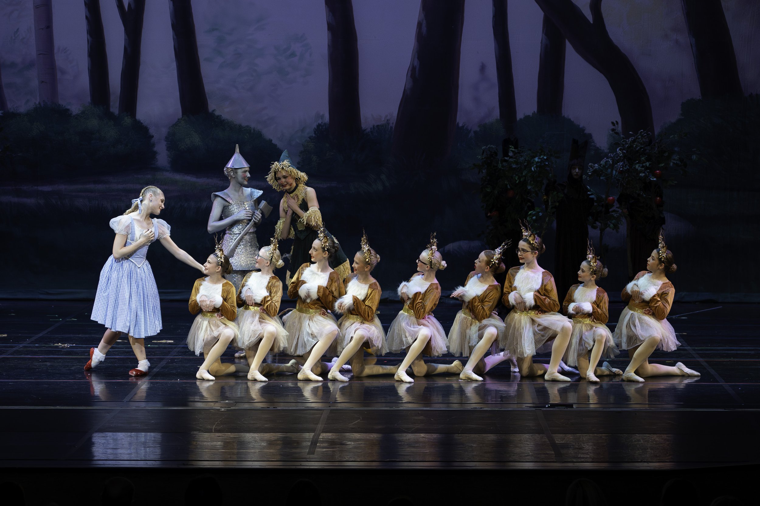 Children dressed as mice performing a ballet on stage, with characters from The Wizard of Oz, including the Tin Man and the Wizard, in a forest-themed backdrop.