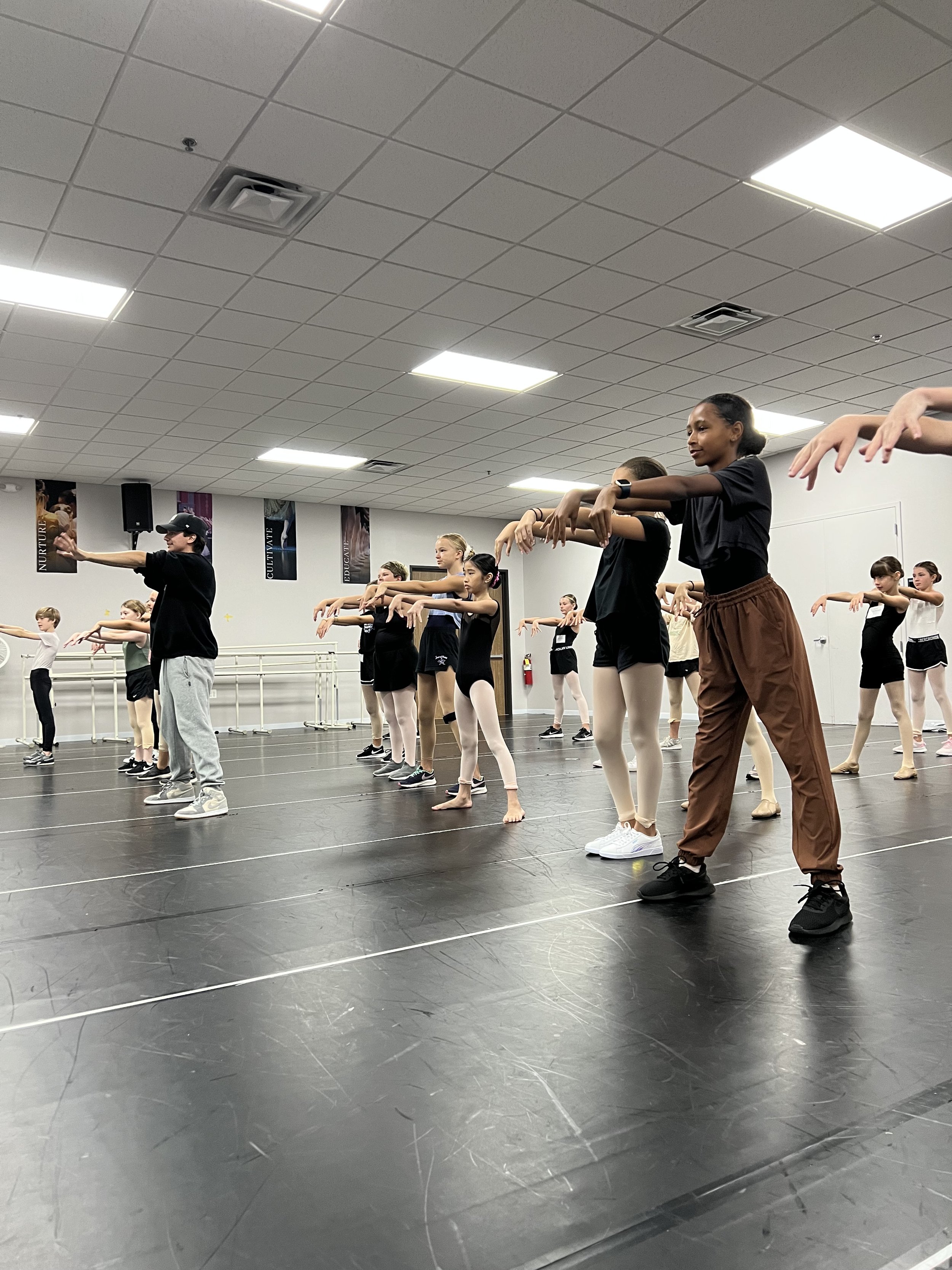 Group of young ballet dancers practicing at a dance studio, following dance instructor in black cap and gray sweatpants. Dancers wearing ballet attire, some in leotards and tights, are standing on a black dance floor with ballet bars along the walls.