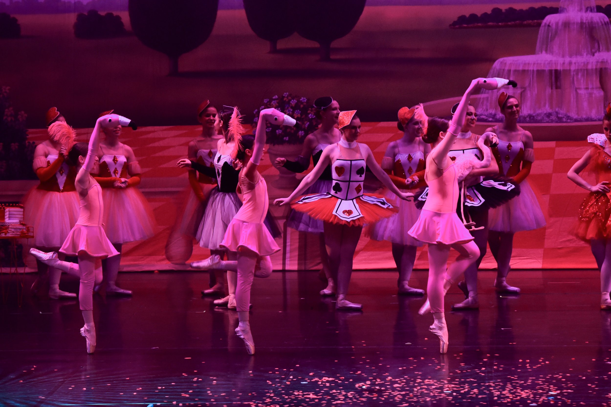 Ballet dancers dressed as playing cards and flamingos performing on stage with a painted backdrop of a garden and fountain.