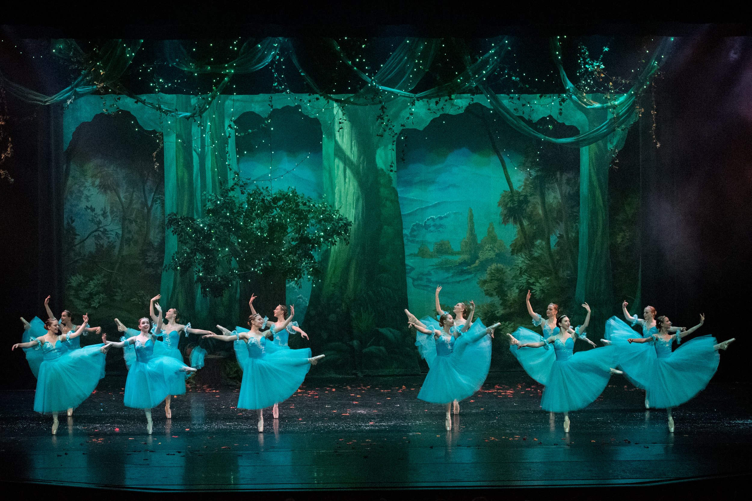 Ballet dancers in blue costumes performing on stage with a forest-themed backdrop and green lighting.