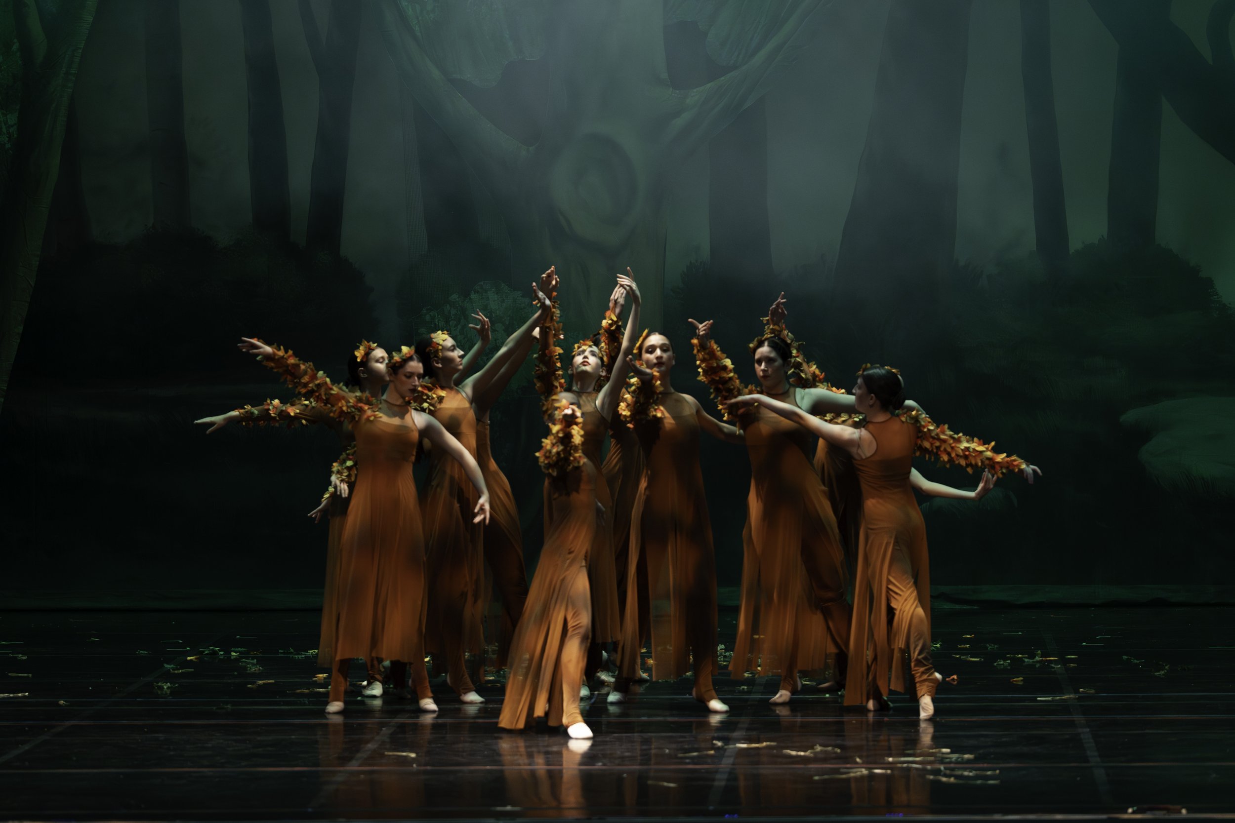 Group of dancers dressed in orange costumes with fall leaves, performing on a stage with a forest backdrop.