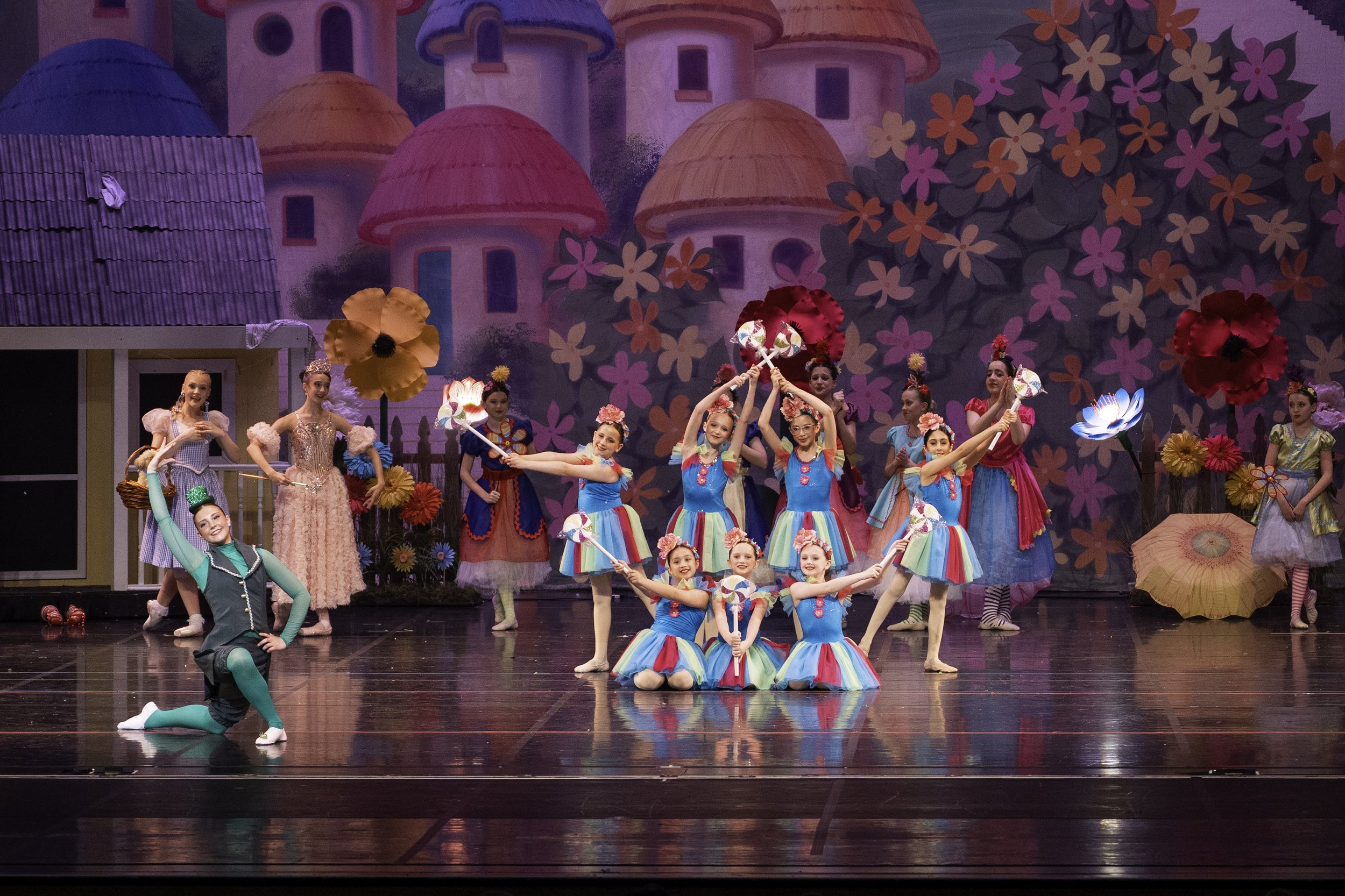 A group of young girls in colorful costumes performing a dance on stage with a whimsical, fairy tale castle backdrop, large flowers, and props.