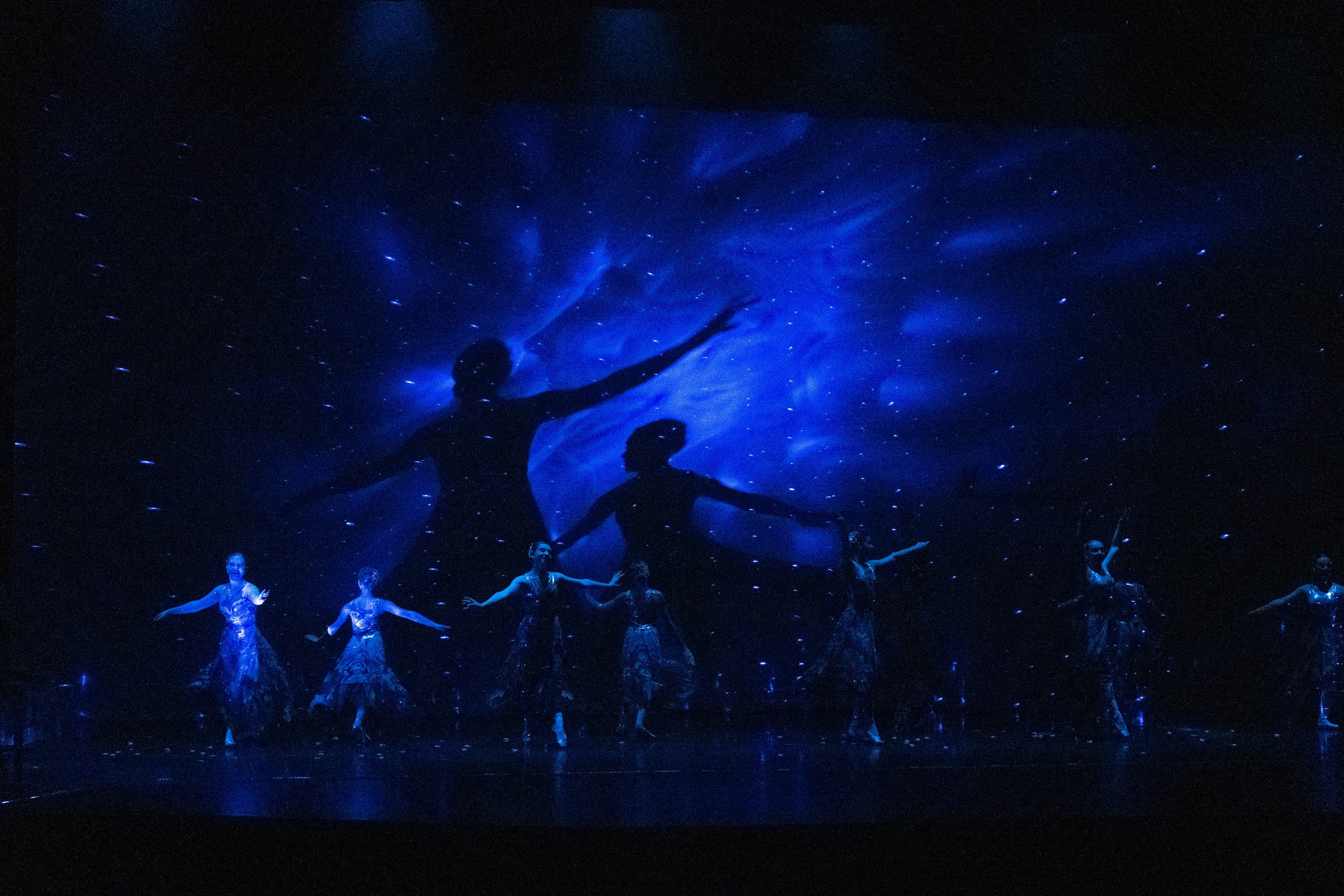 A dance performance with a backdrop of a starry night sky and two large silhouettes of dancers with arms outstretched, illuminated in blue light.